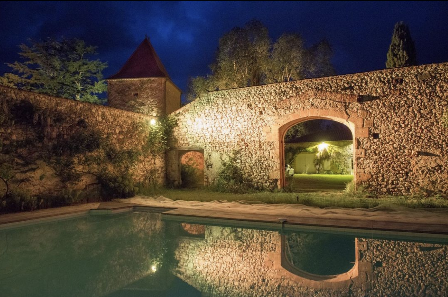 Piscine à l'extérieur d'une vieille structure en pierre, avec un mur en pierre et une tour avec toit en forme de pyramide, éclairée la nuit, reflet dans l'eau de la piscine.