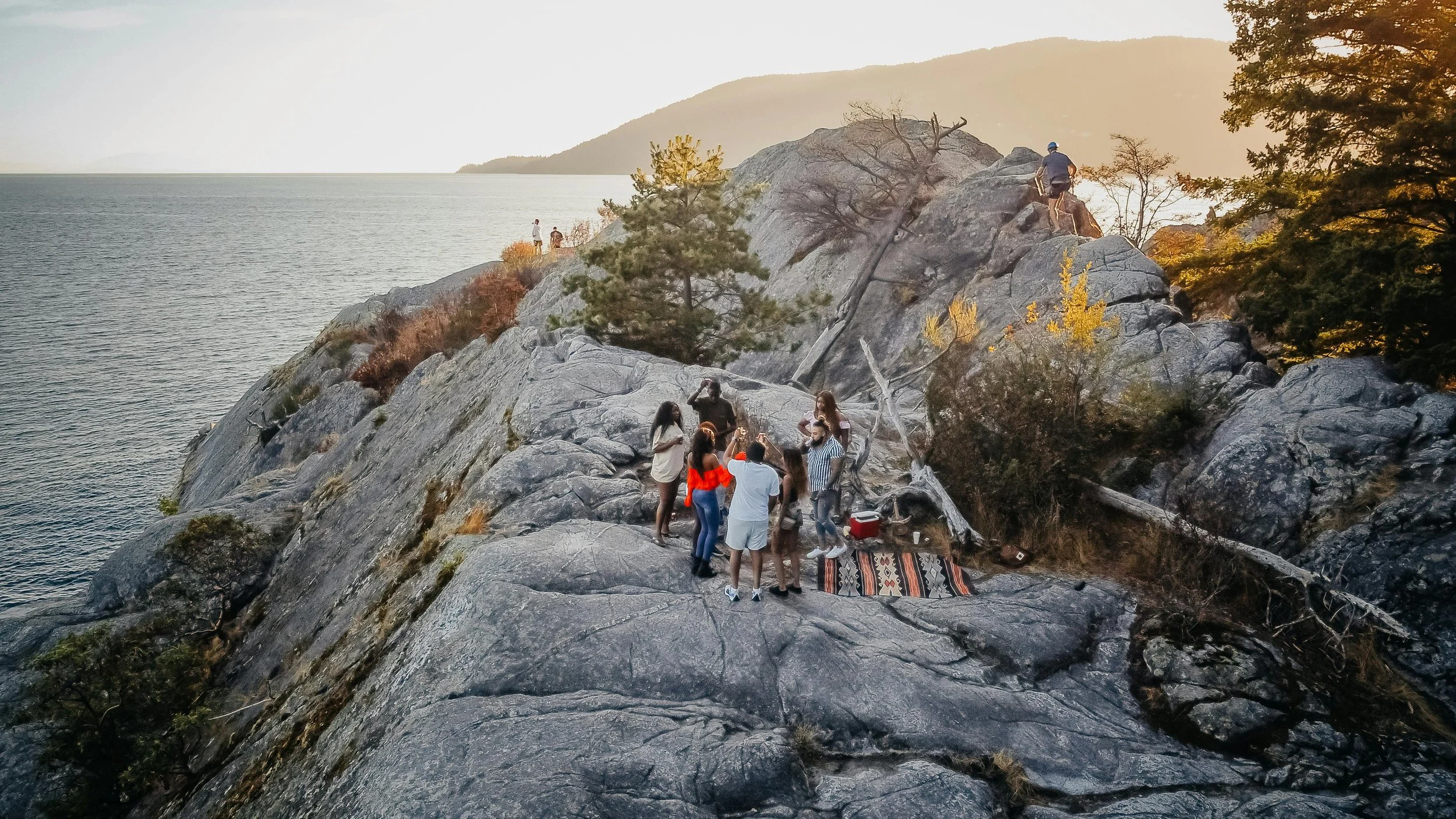 Groupe de personnes faisant un pique-nique et prenant des photos sur des rochers en bord de lac ou de mer au coucher du soleil.