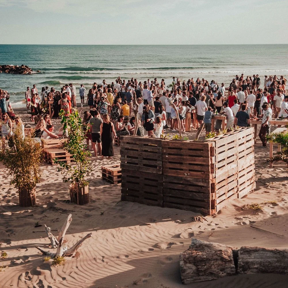 Une foule de personnes sur une plage, certains à l'eau, d'autres autour de tables et de palissades en bois, avec la mer en arrière-plan et une atmosphère ensoleillée.