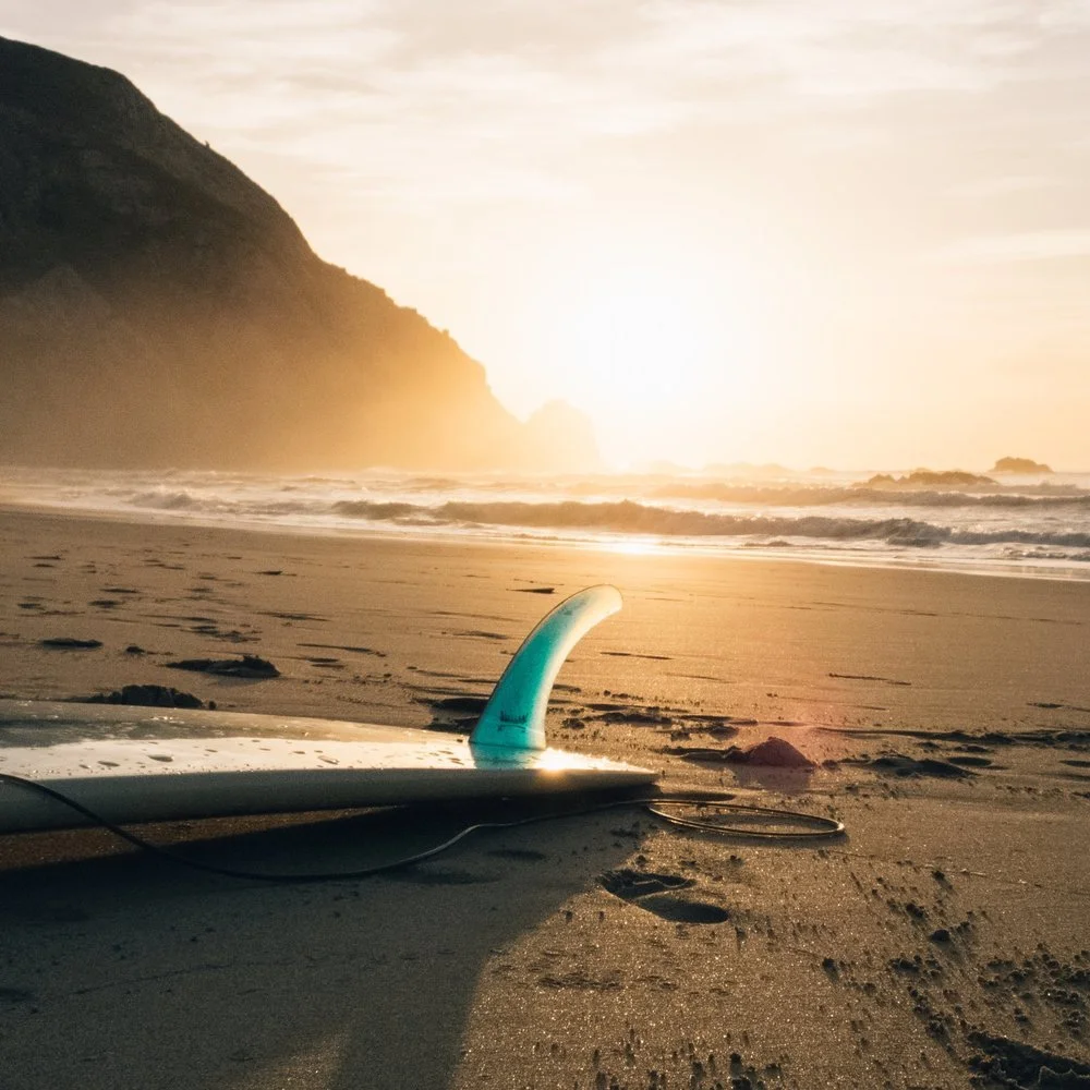 Planche de surf avec quille bleu turquoise sur le sable de la plage de Biarritz au coucher du soleil, avec des vagues déferlantes et une falaise en fond.
