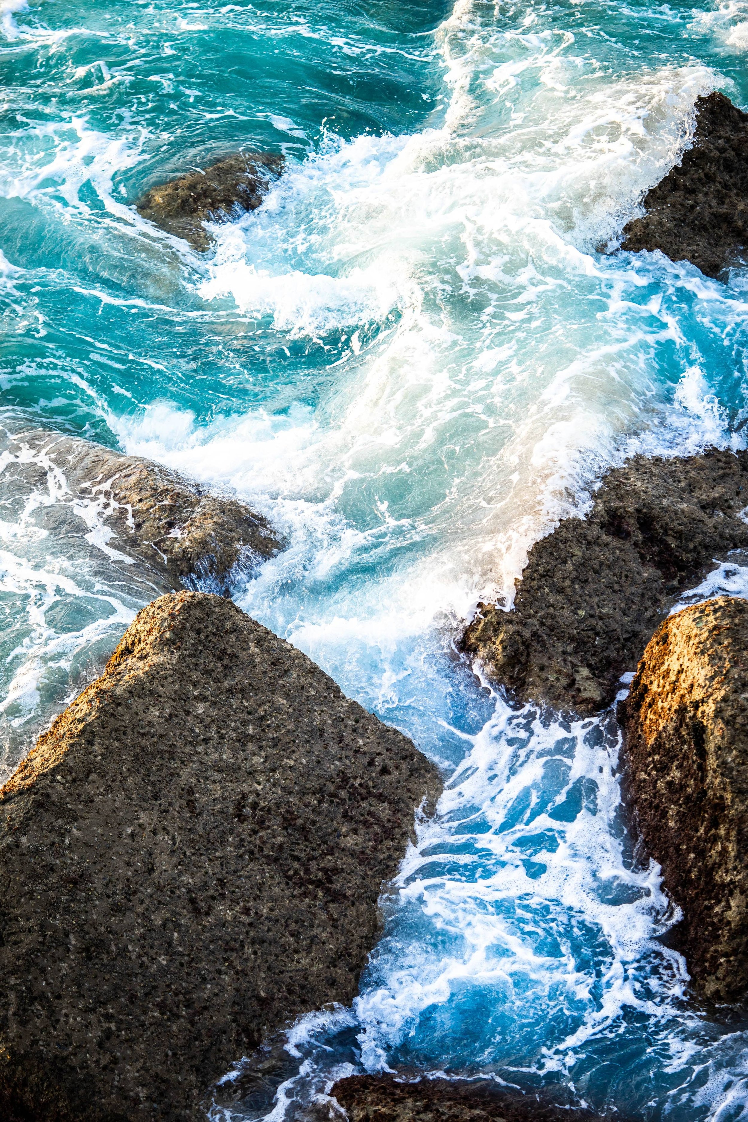 Gros plan des vagues turquoises se brisant avec de la mousse blanche sur les rochers sombres sur la côte, mettant en évidence la force et le mouvement de l’océan
