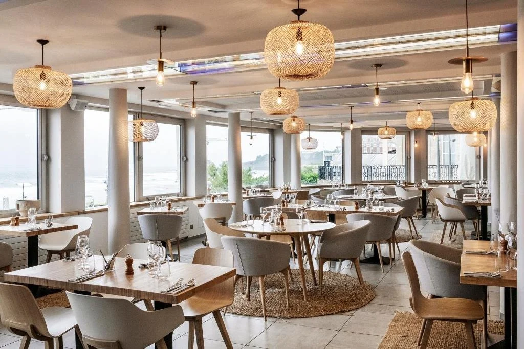 Restaurant de l’Hôtel Windsor Biarritz avec un design moderne, lampes en osier et grandes fenêtres donnant sur la mer, offrant une ambiance lumineuse pour les convives.