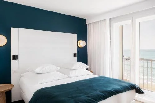 Chambre d’hôtel à l’Hôtel Windsor Biarritz avec un design côtier moderne, des murs bleu marine et blanc, et de grandes fenêtres. Grand lit avec tête de lit blanche.
