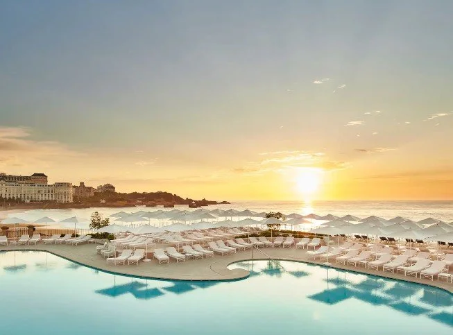 Piscine extérieure de l’Hôtel du Palais avec transats prêts, baignée par la lumière dorée du coucher de soleil sur l’océan, offrant une vue panoramique spectaculaire.