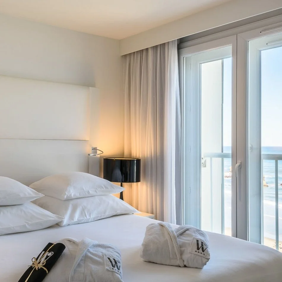 Chambre d’hôtel avec un design moderne et minimaliste. Un grand lit et des draps blancs, avec des peignoirs de courtoisie pliés sur elle, à côté d’un balcon.