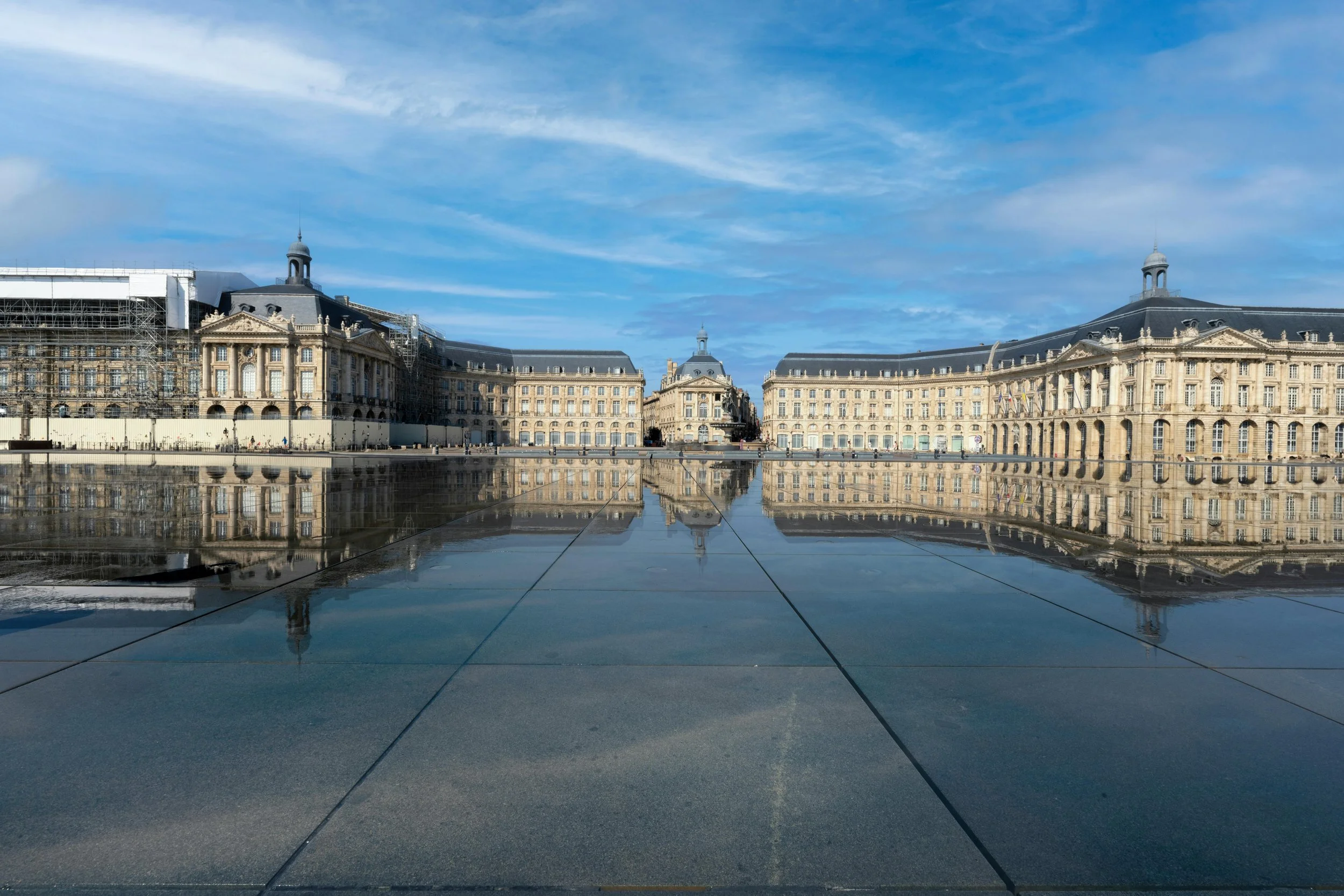 Vue de la place historique de la Bourse à Bordeaux, avec ses bâtiments classiques se reflétant dans le miroir d’eau (Miroir d’eau) sous un ciel bleu.