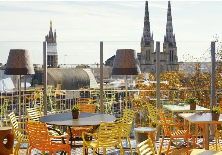 Terrasse sur le toit du Mama Shelter Bordeaux avec tables et chaises, offrant une vue panoramique sur les toits et les volutes historiques de la ville, idéal pour les loisirs urbains.