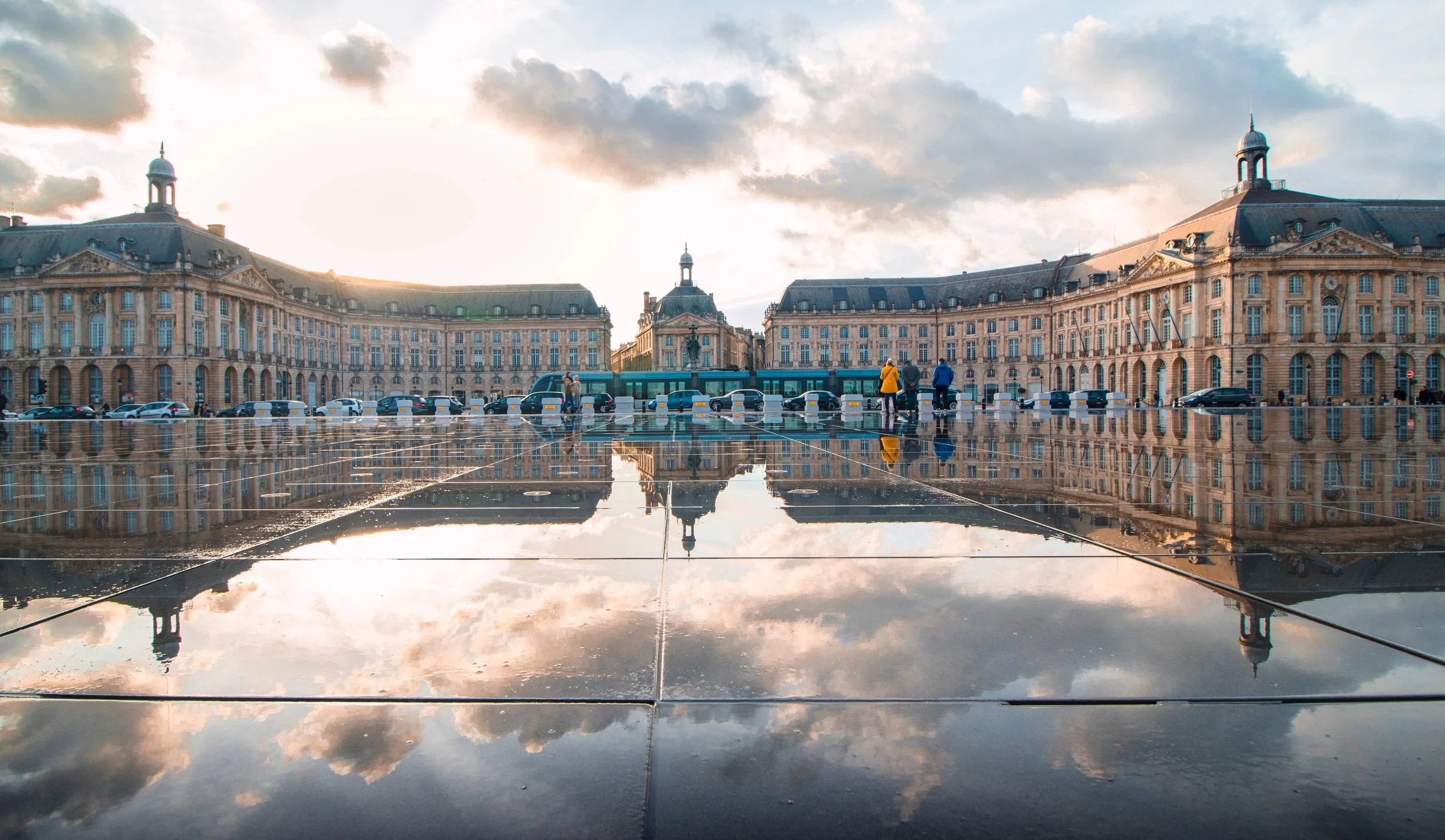 Vue de bâtiments historiques avec un ciel nuageux, reflet dans une grande surface d'eau ou de mall. Personnes marchant près d'un bus.