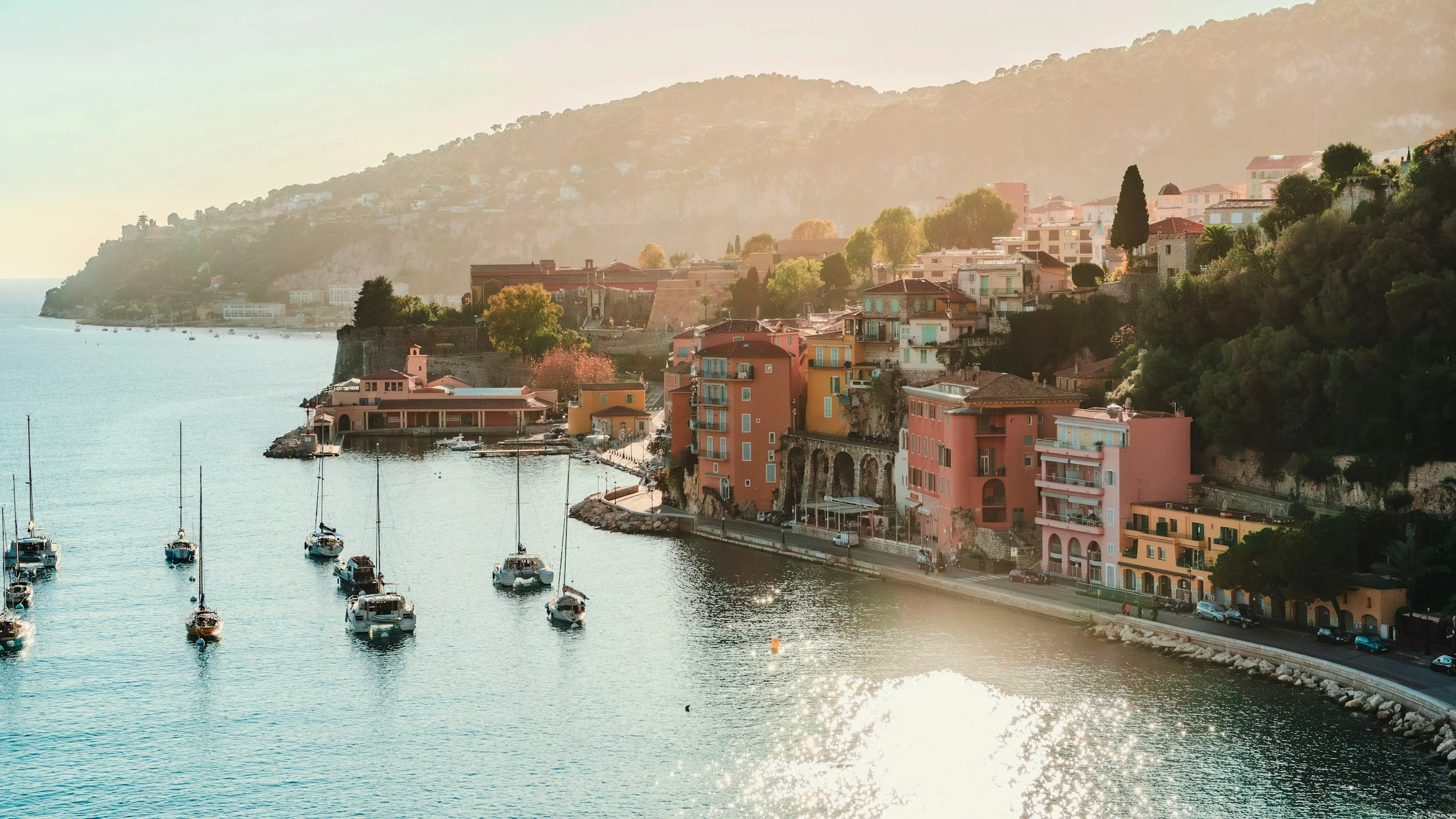 Vue d'un port avec des bateaux en mer et des bâtiments colorés en montée le long de la côte, avec des montagnes en arrière-plan sous une lumière douce.