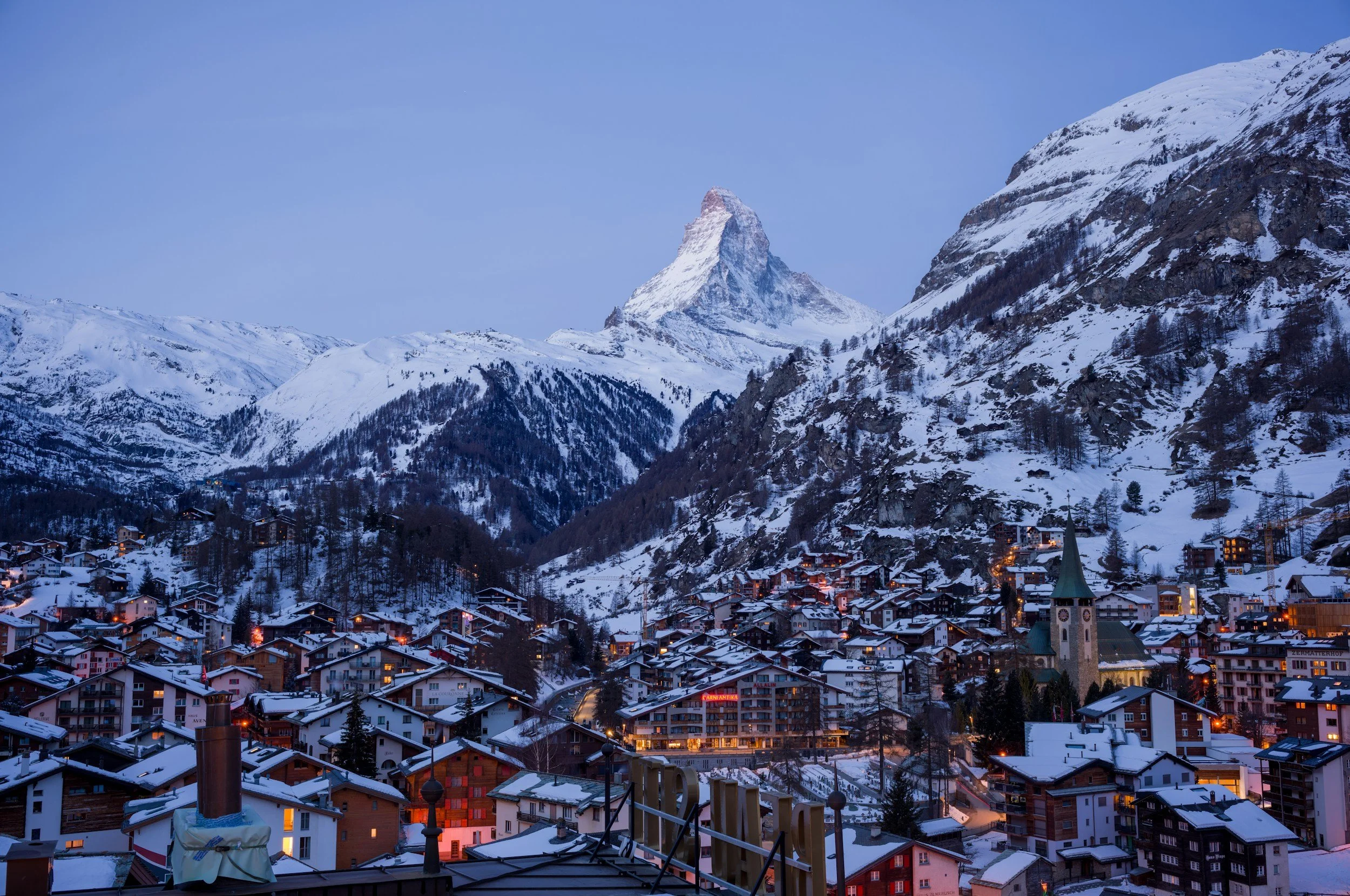 Vue panoramique d'une ville alpine enneigée avec une montagne emblématique, le Cervin, en arrière-plan, et des maisons illuminées dans la vallée.