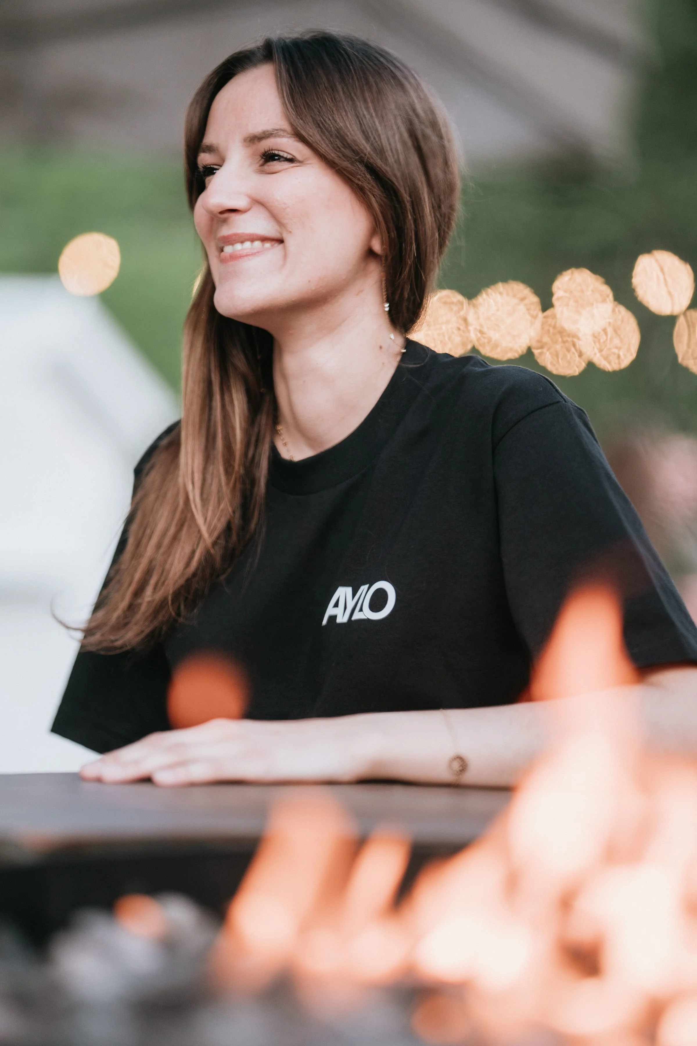 Une femme souriante avec des cheveux bruns longs, portant un t-shirt noir avec le logo 'AMLO', assise à une table, avec un fond flou de lumières chaudes.