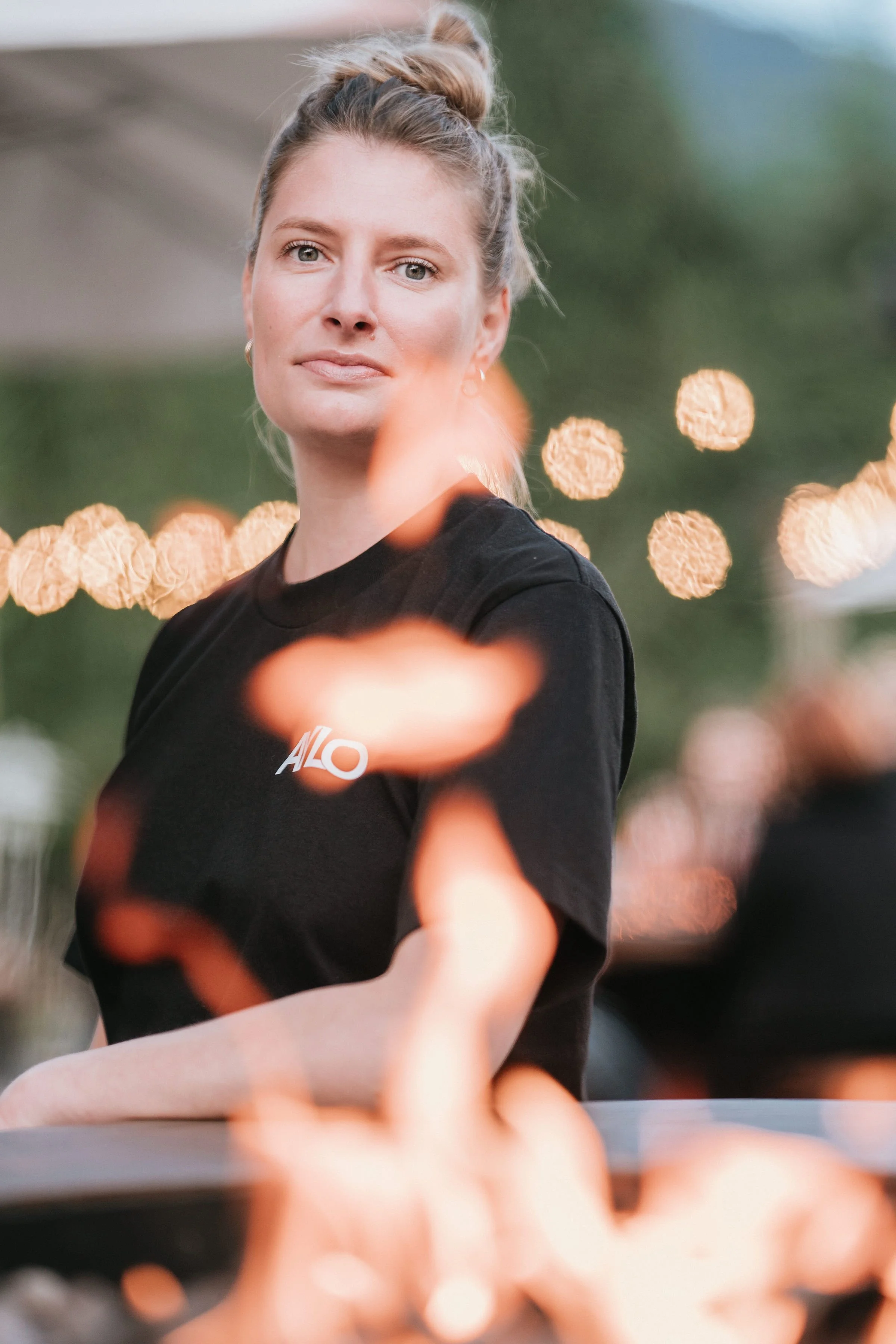 Une femme avec des cheveux blonds attachés, portant un t-shirt noir avec un logo blanc 'ALO', se tenant à l'extérieur, avec une lumière douce en arrière-plan.