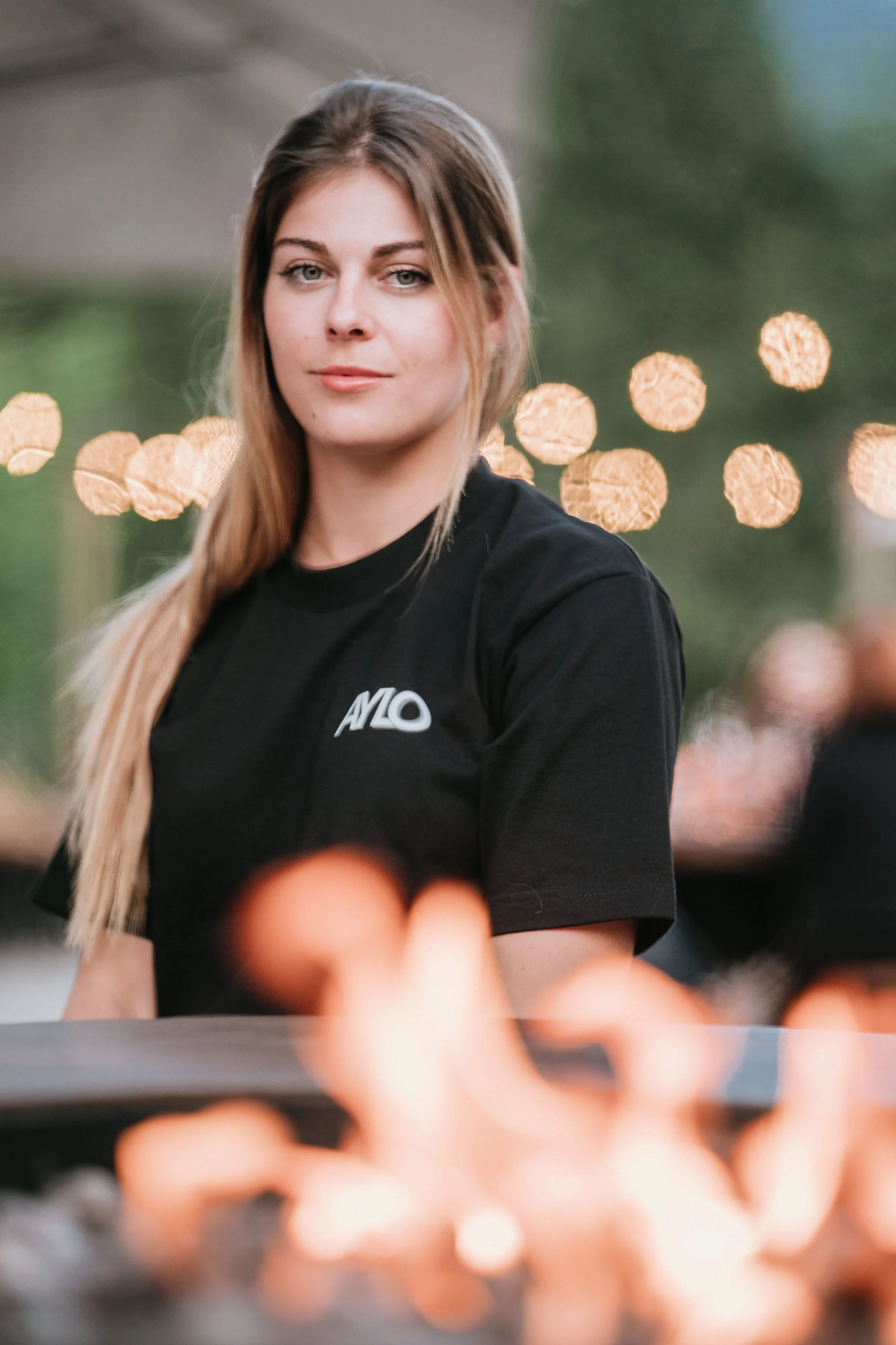 Une jeune femme avec des cheveux longs, portant un t-shirt noir avec un logo « AYLO » à l'avant, regardant vers l'appareil photo, avec un arrière-plan flou de lumières de ville ou de décor nocturne.