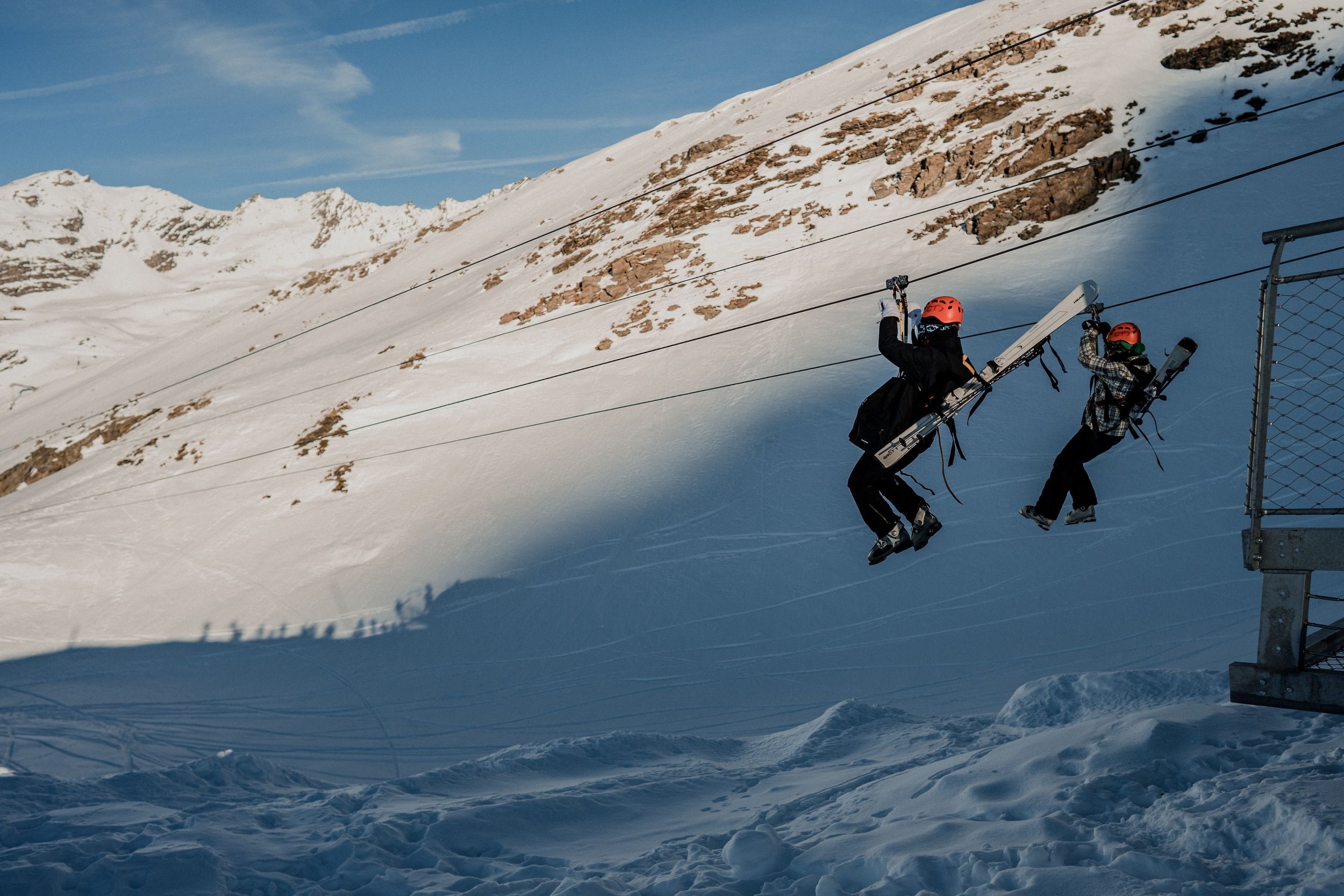 Deux skieurs en train de monter une pente enneigée à l'aide d'un télésiège dans un paysage de montagnes rocheuses et enneigées sous un ciel bleu.
