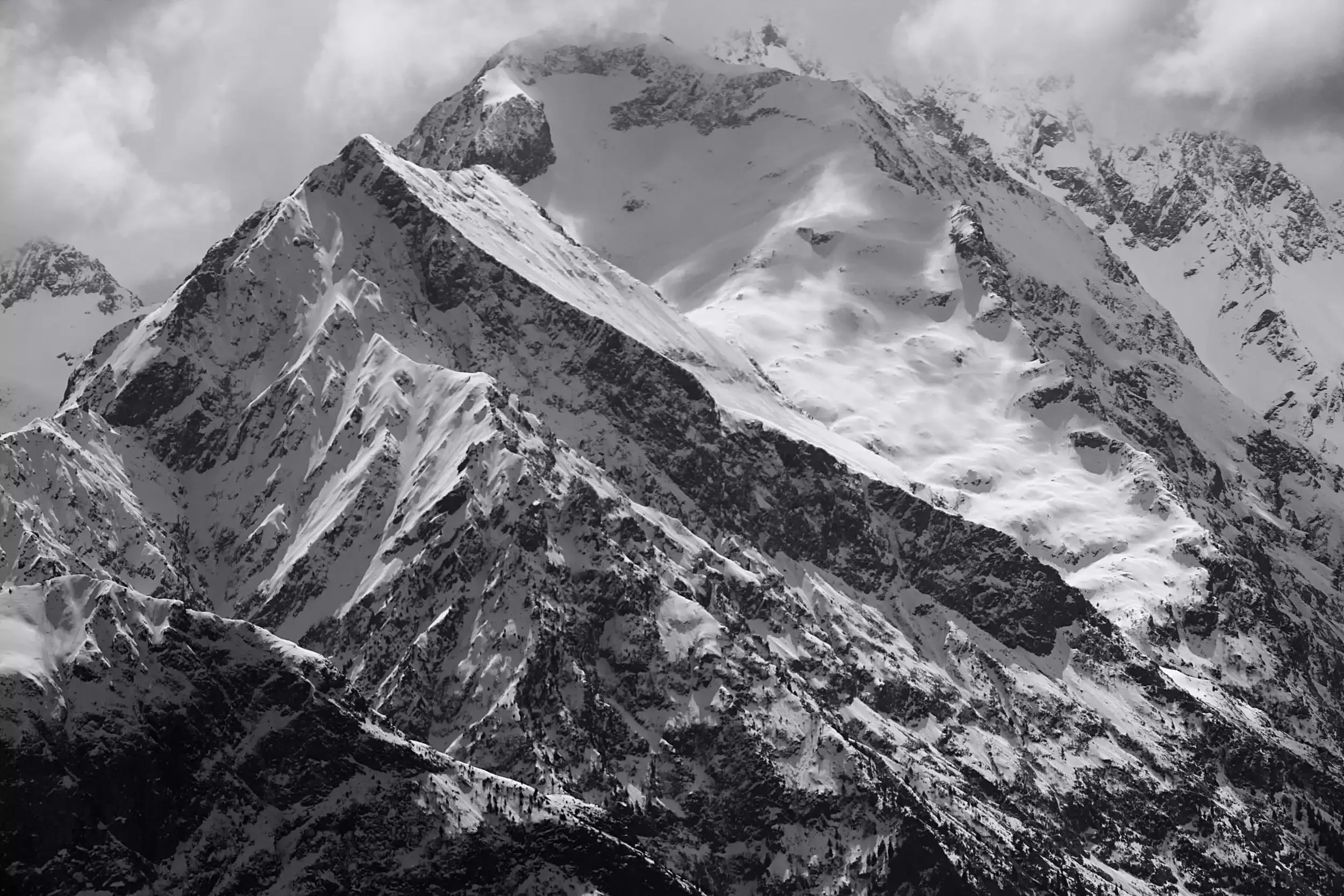 Une montagne imposante des Alpes, avec ses pentes escarpées et couvertes de neige, photographiée en noir et blanc. Dans la partie basse, on peut observer des forêts de pins et un ciel nuageux.