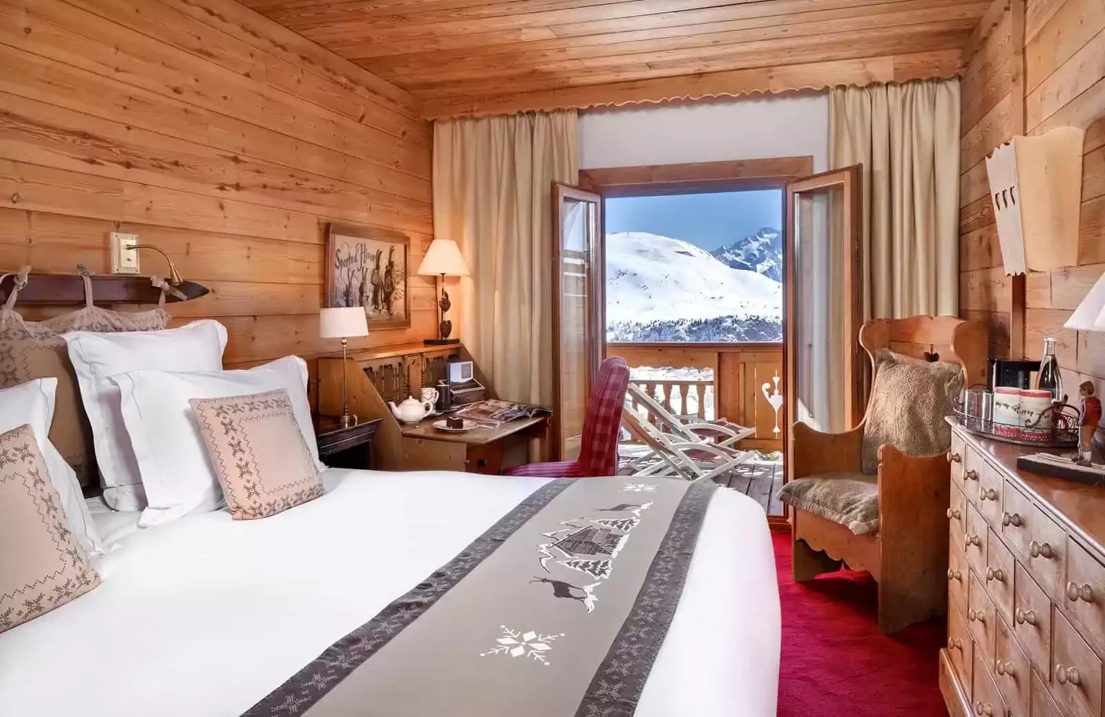 Chambre de style chalet à l'Hôtel Chamois d'Or 5* à L'Alpe d'Huez, avec boiseries, grand lit et balcon ouvert offrant une vue montagneuse. Confortable pour un séminaire Aylo Montagne.