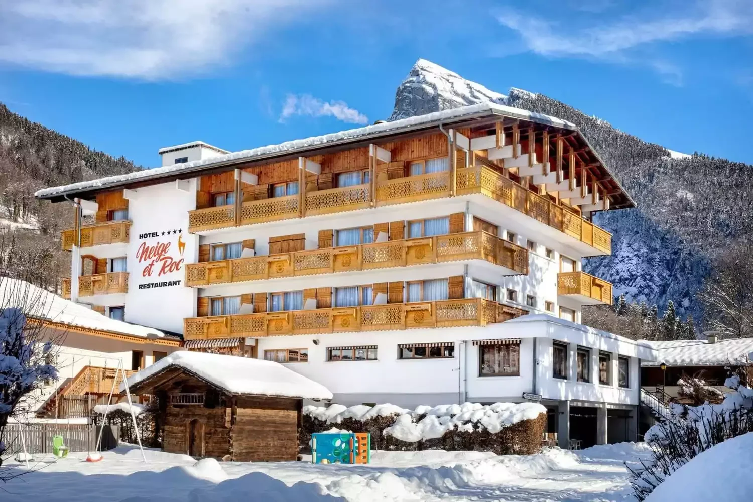 Façade du Hôtel Neige et Roc 4*, un hôtel de montagne de style chalet avec balcons en bois. Le bâtiment est couvert de neige et dispose d’une aire de jeux enneigée à l’extérieur. En arrière-plan, on voit des montagnes alpines majestueuses.