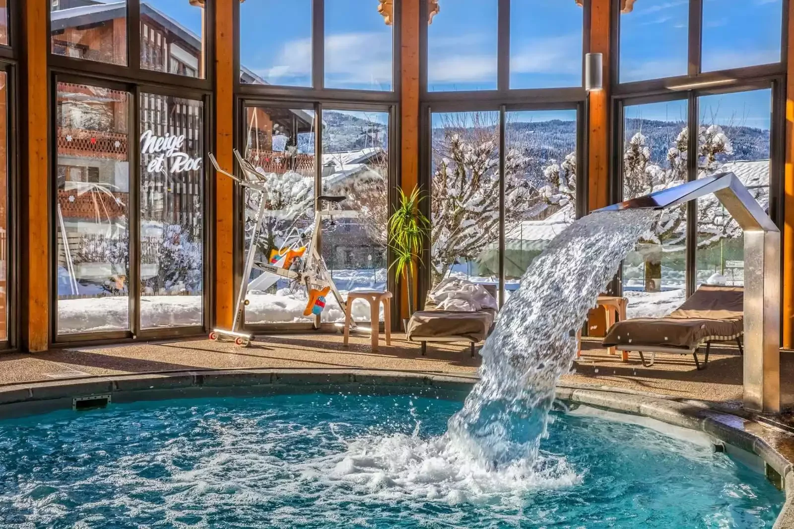 Piscine intérieure chauffée au **Hôtel Neige et Roc ****, avec de grandes baies vitrées offrant une vue panoramique sur les montagnes enneigées et un jet d’eau qui tombe dans la piscine.