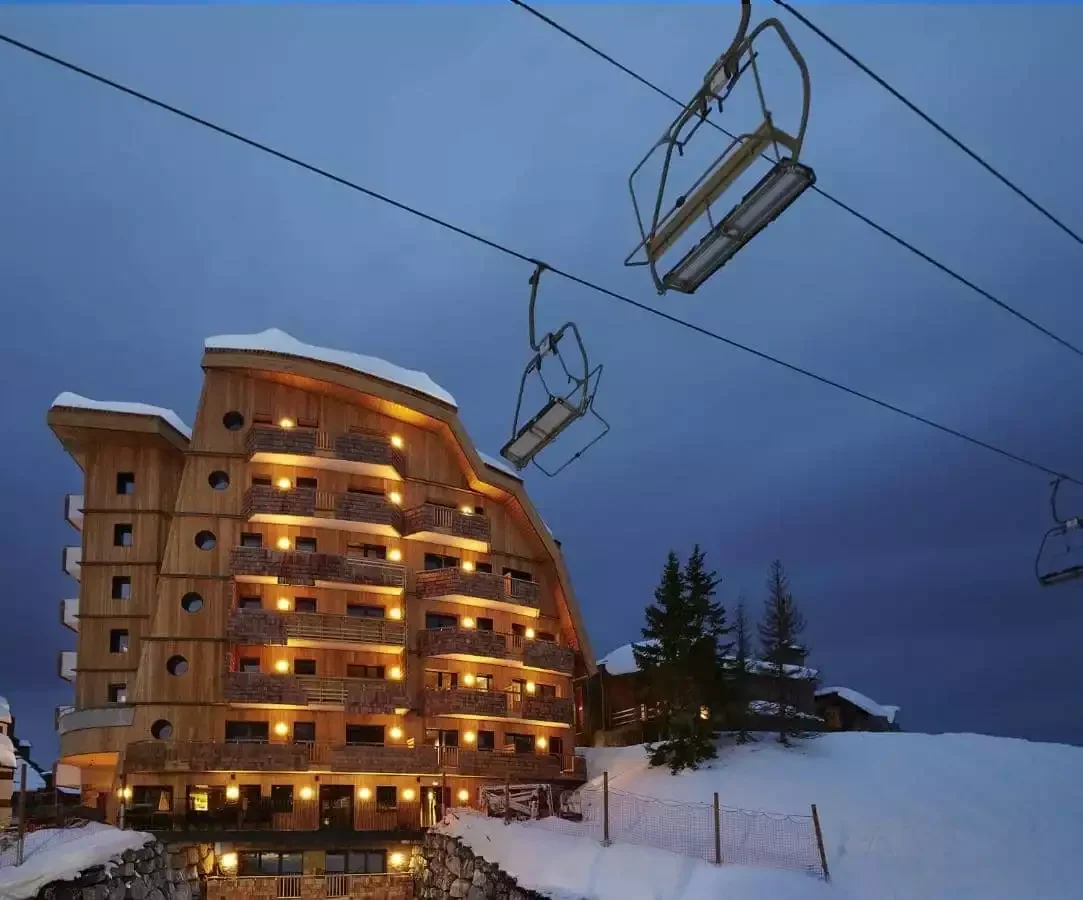 Vue nocturne de l’Hôtel Mil8 4*, un chalet à l’architecture moderne et façade en bois, éclairé sous un ciel sombre. Un télésiège pend au-dessus, indiquant sa proximité des pistes