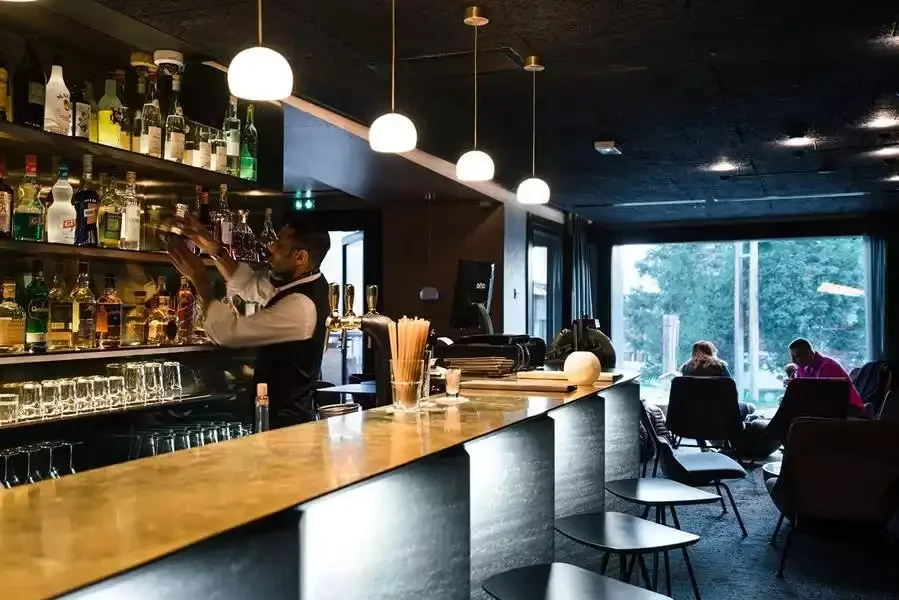 Bar moderne à l’Hôtel Mil8 4* avec un comptoir en bois éclairé et un barman préparant des boissons. L’espace offre une atmosphère accueillante pour se détendre.