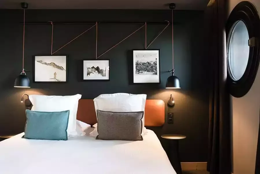 Chambre design à l’Hôtel Mil8 4*, avec un grand lit, une décoration minimaliste avec des murs sombres et des éléments artistiques, et une fenêtre ronde, offrant une ambiance moderne et élégante.