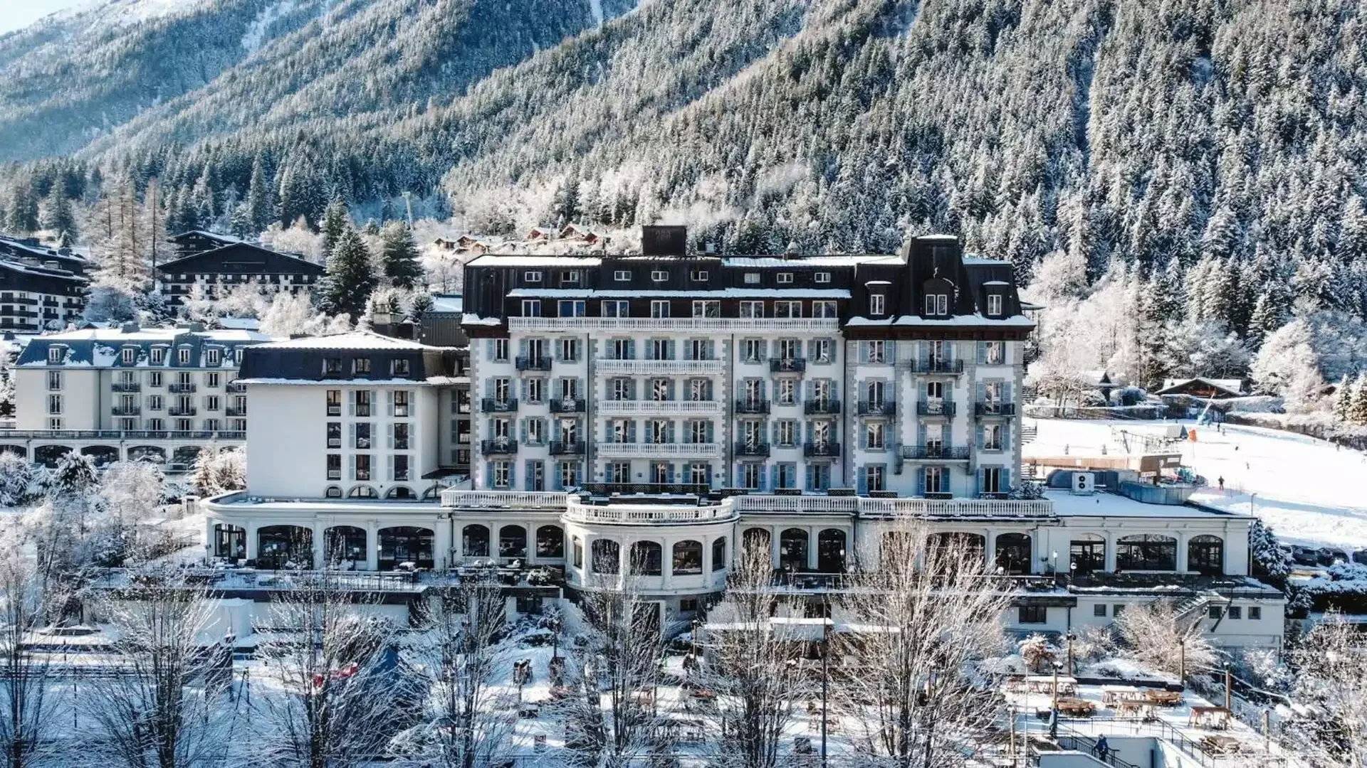 Façade extérieure de l’Hôtel La Folie Douce 4* à Chamonix, un grand bâtiment de style classique avec balcons, toits enneigés et entouré d’un paysage hivernal et de montagnes boisées.
