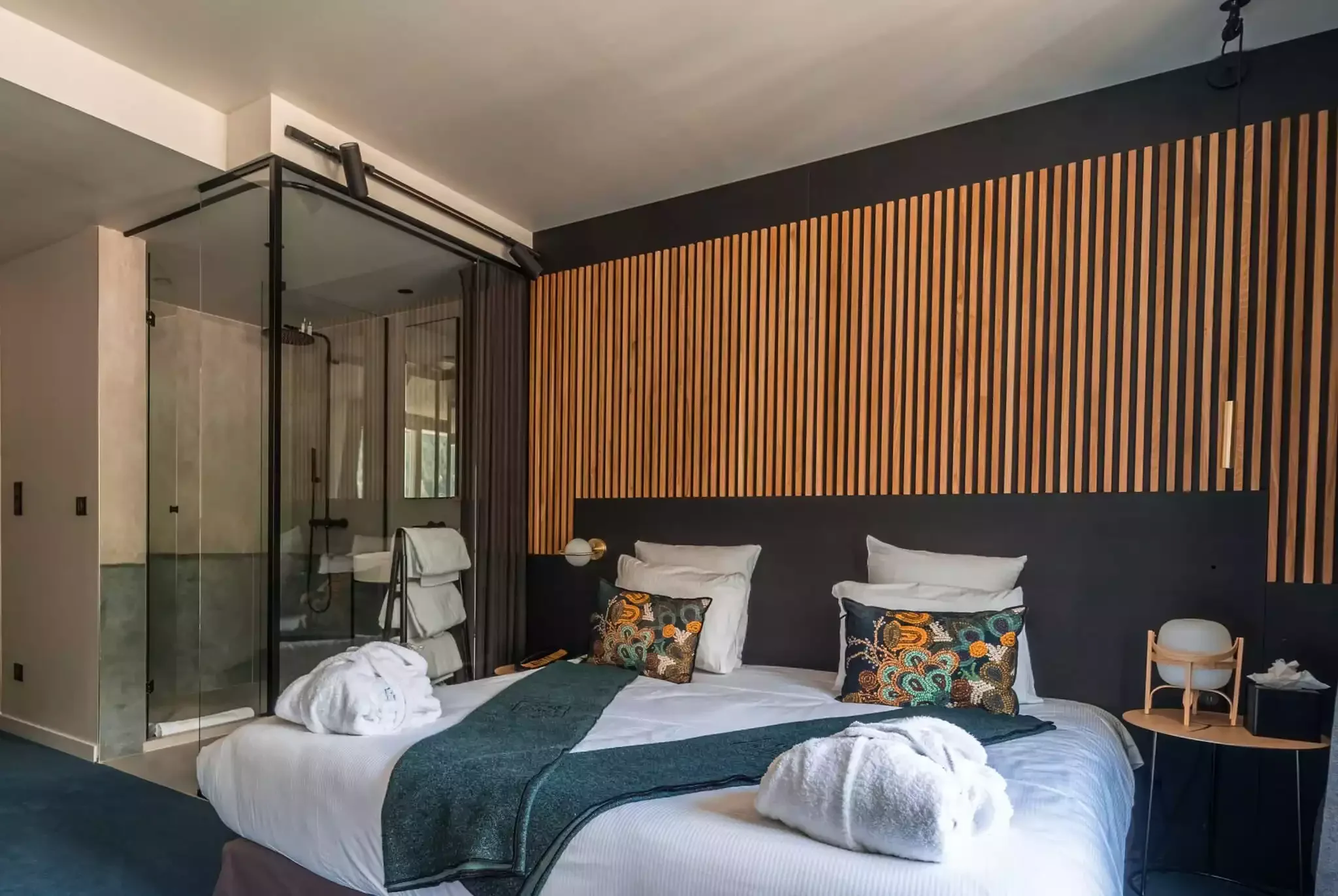 Chambre moderne à l’Hôtel La Folie Douce 4* avec un grand lit, une tête de lit en bois, des murs sombres et une salle de bain en verre transparent, créant un design élégant.