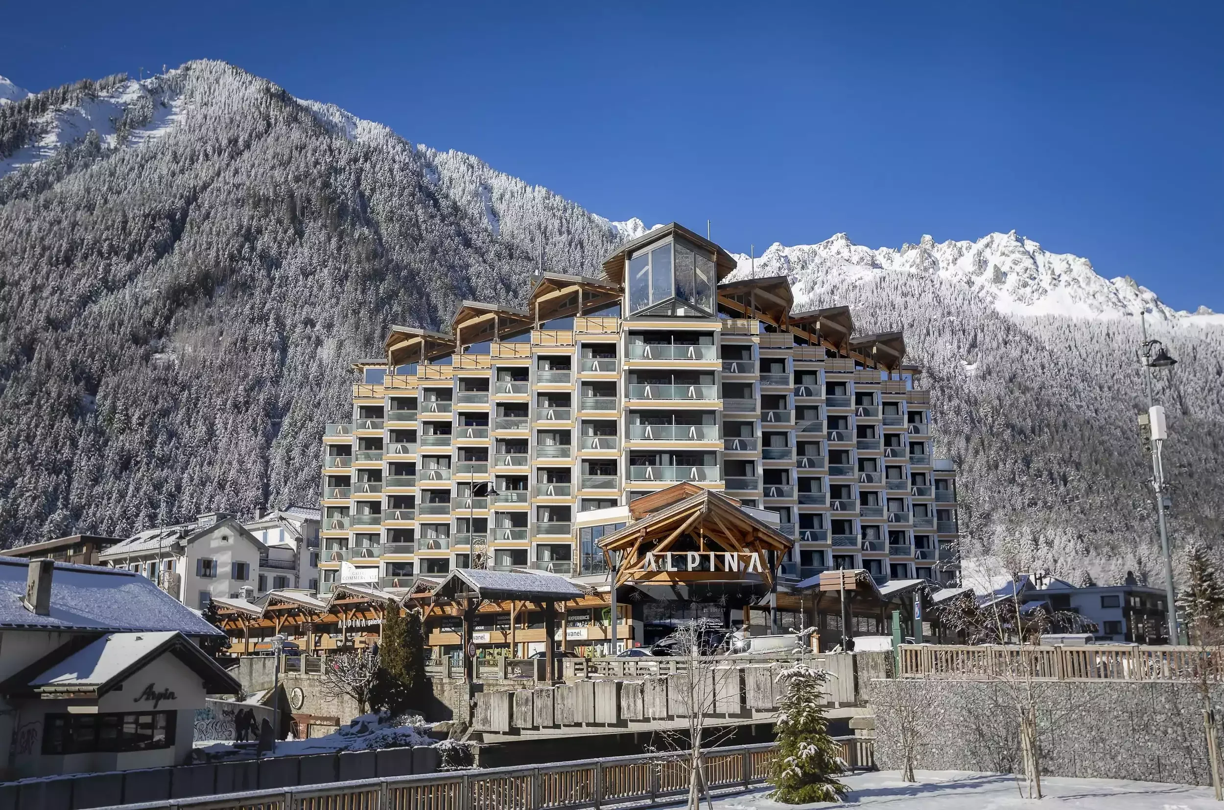 Façade moderne de l’hôtel Alpina, un imposant bâtiment de montagne avec une architecture unique et des balcons en bois. Il est situé dans un village enneigé avec des pics alpins majestueux à l’arrière-plan.
