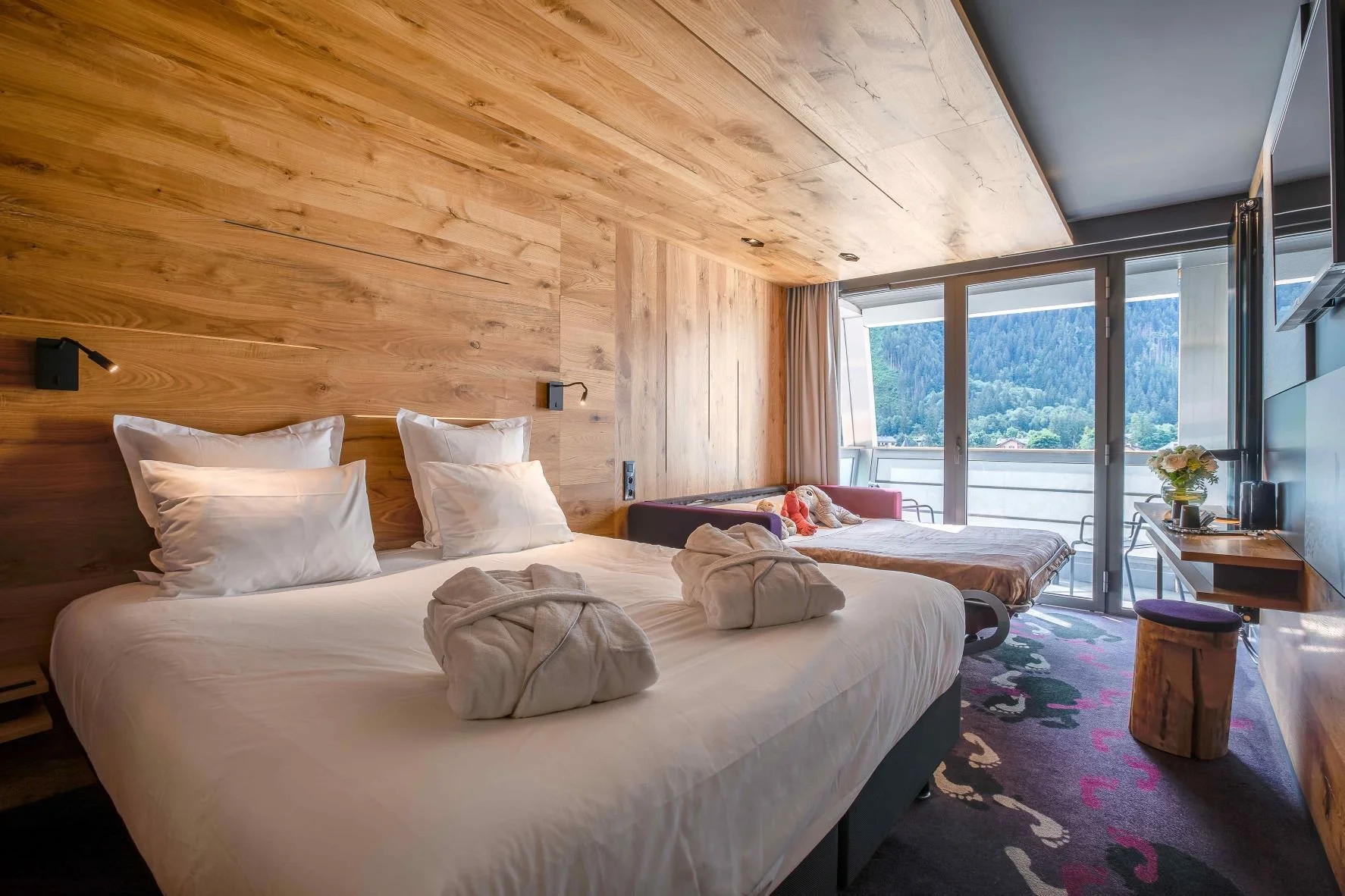 Chambre design à l’Hôtel Alpina Chamonix, avec des murs en bois clair et un grand lit, qui s’ouvre sur un balcon avec vue imprenable sur les montagnes.