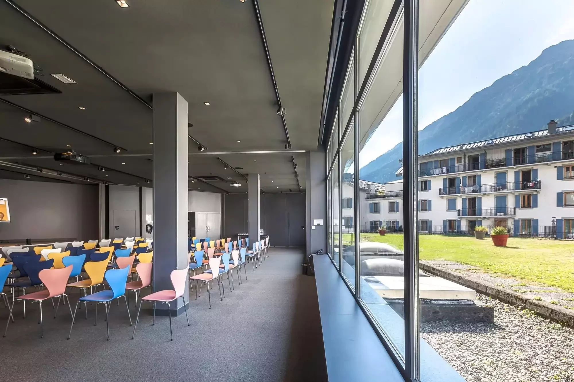 Salle de réunion de l’Hôtel Alpina Chamonix, équipée de chaises colorées et de grandes fenêtres donnant sur un jardin extérieur, idéale pour des événements d’entreprise ou des ateliers.