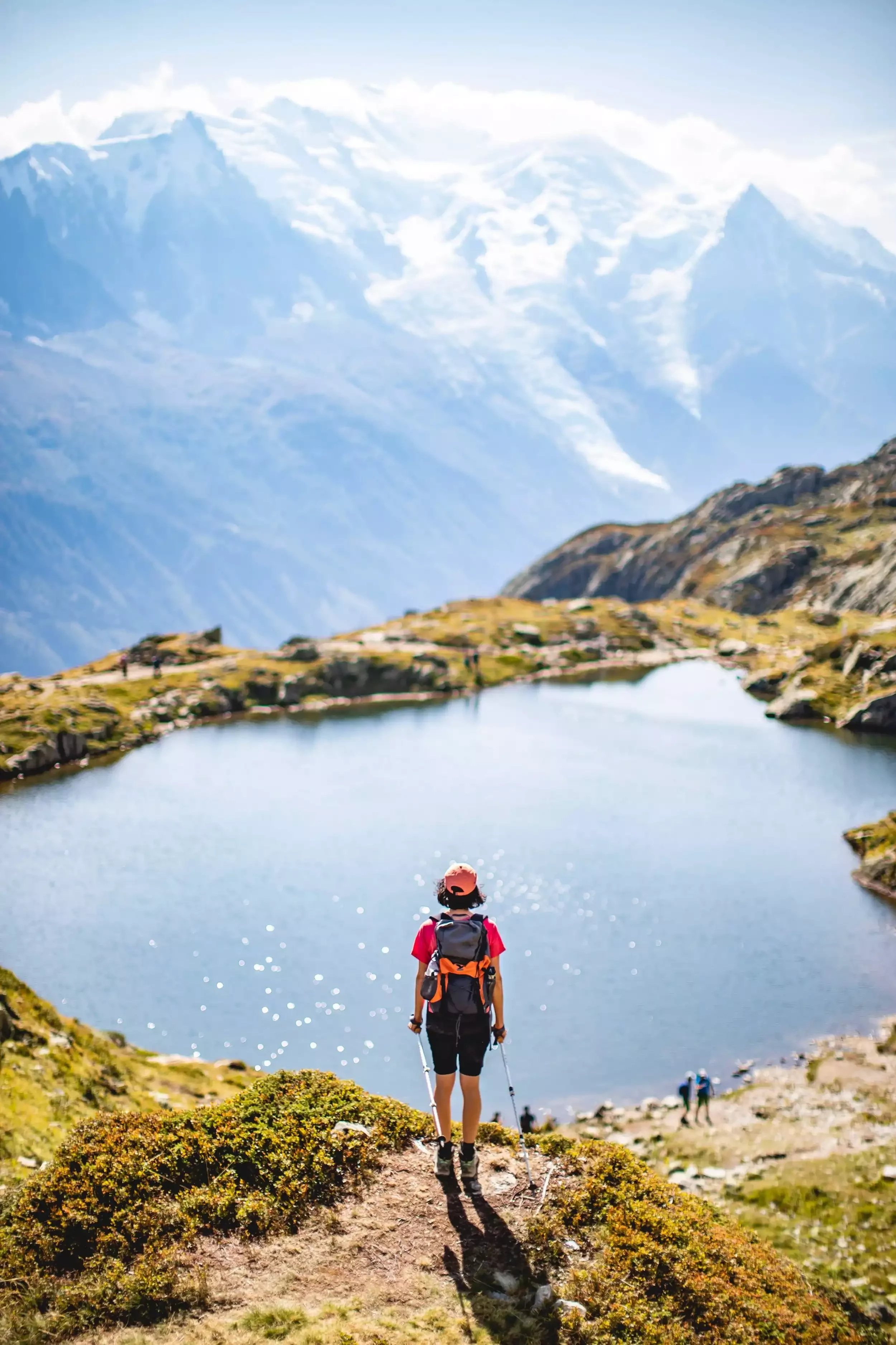 Un participant à un séminaire Aylo Montagne pratique la randonnée avec son sac à dos et ses bâtons, en s’approchant d’un lac de montagne à Chamonix Été. La vue ensoleillée des Alpes offre un cadre idéal pour les activités d’équipe