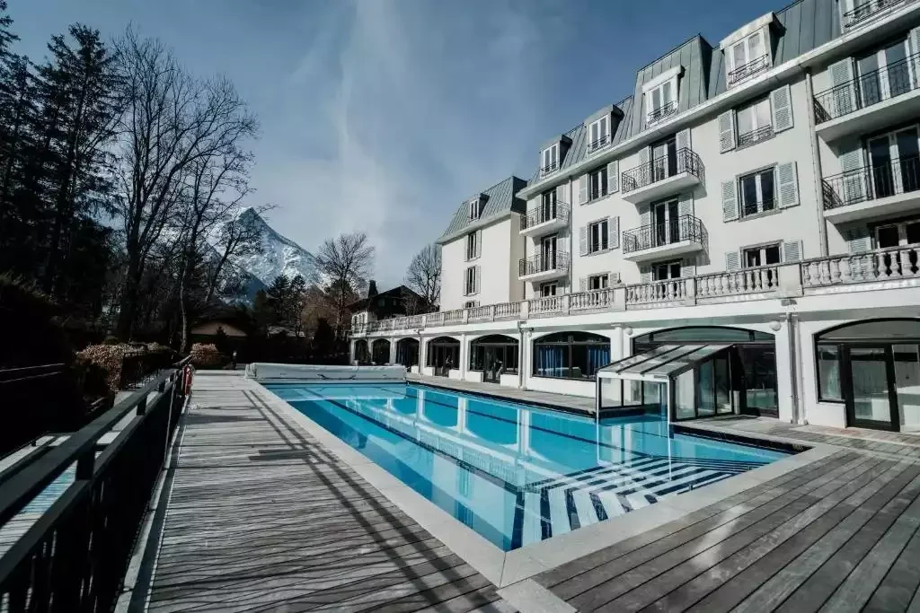 Piscine extérieure de l’Hôtel La Folie Douce 4* à Chamonix Été, avec une zone couverte et un solarium en bois, bordée par la façade classique de l’hôtel et la verdure.