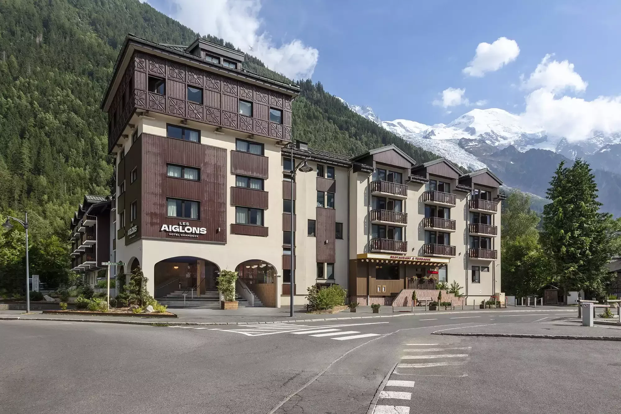Vue extérieure de l’Hôtel Les Aiglons à Chamonix, un bâtiment de style chalet moderne avec des détails en bois et balcons. Il est situé dans une rue de la ville, avec un paysage de montagnes verdoyantes et des sommets enneigés en arrière-plan.