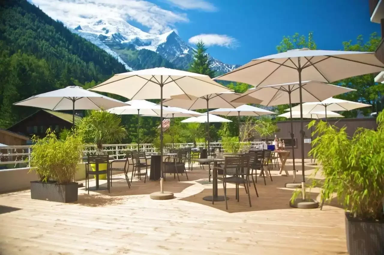 Terrasse extérieure de l’Hôtel Les Aiglons à Chamonix, avec tables, chaises et grands parasols blancs. Des sommets enneigés se dressent à l’arrière-plan, créant un espace agréable pour se détendre.