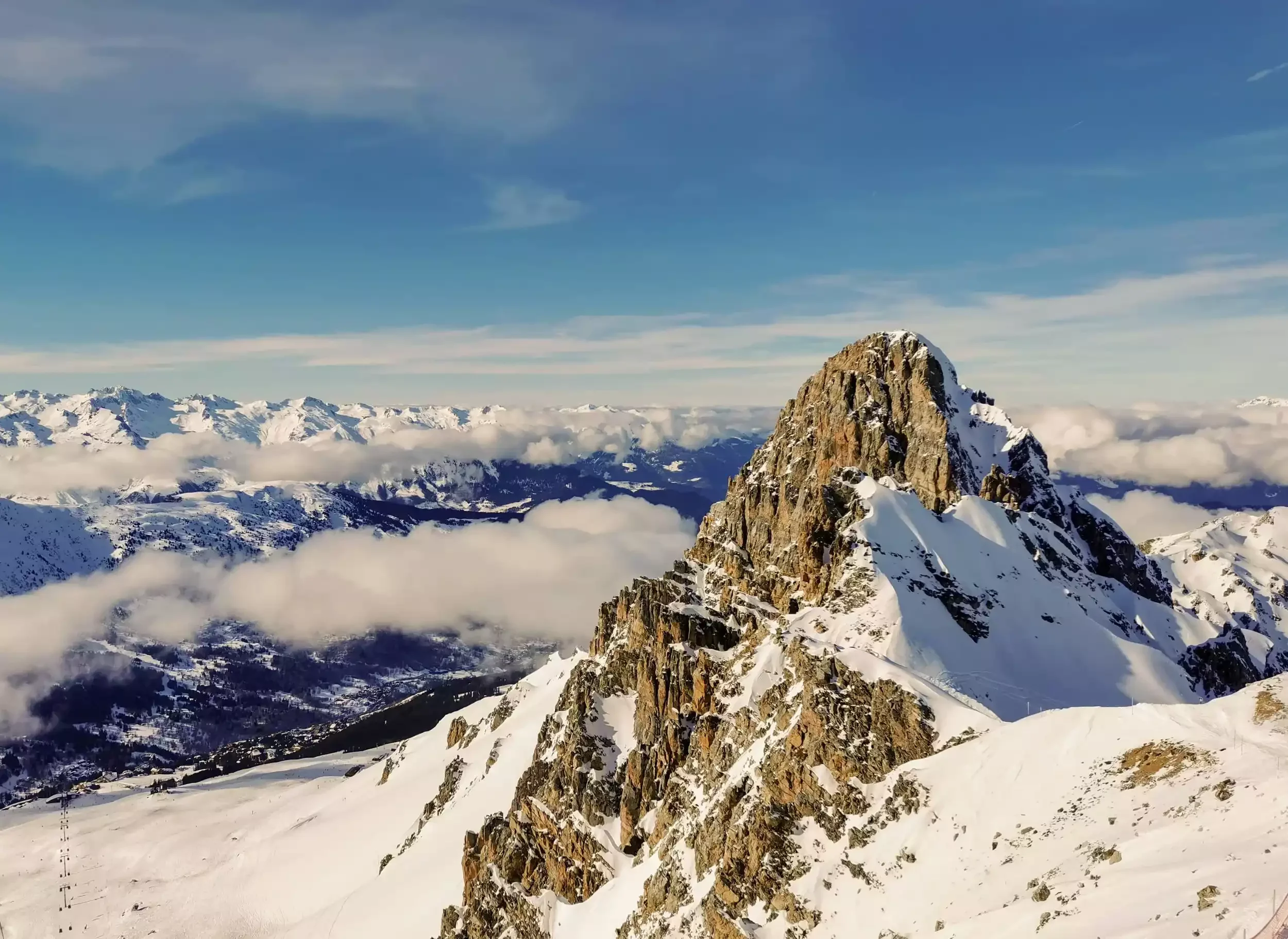Vue panoramique d’un pic alpin rocheux et enneigé à Courchevel, avec une chaîne de montagnes et une mer de nuages recouvrant la vallée. Le ciel bleu avec des nuages dispersés crée un contraste saisissant.
