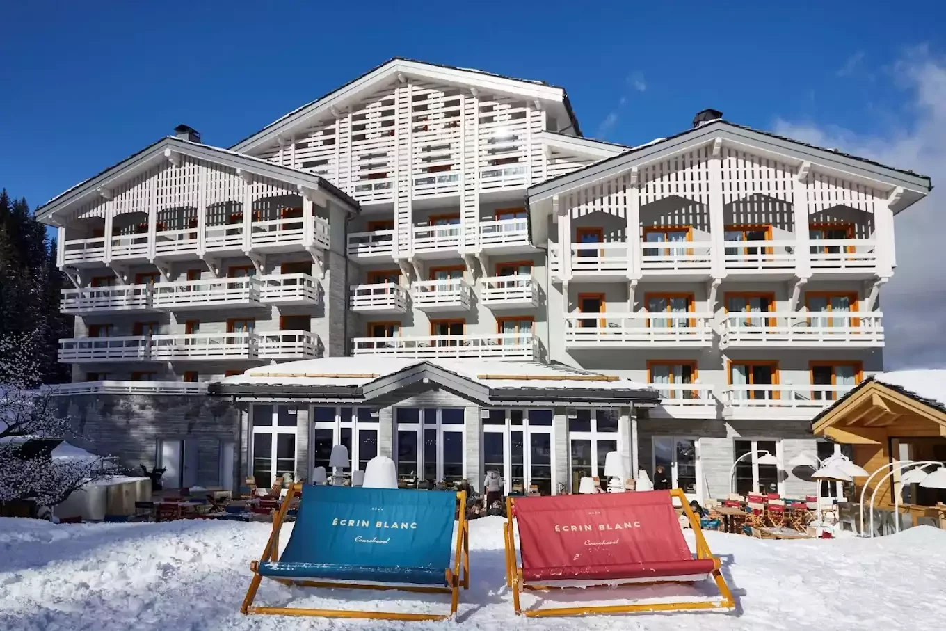 Vue extérieure de l’Hôtel Ecrin Blanc à Courchevel, un grand chalet avec une architecture moderne et des détails en bois blanc. Au premier plan, des chaises de plage bleues et rouges sur la neige, prêtes pour les invités.