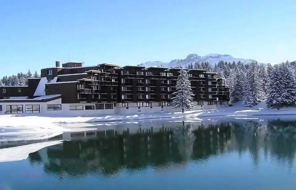 Vue de l’hôtel Lake Courchevel en hiver, un grand bâtiment de style chalet reflété dans un lac de montagne, avec des pins enneigés et majestueuses montagnes alpines en arrière-plan.