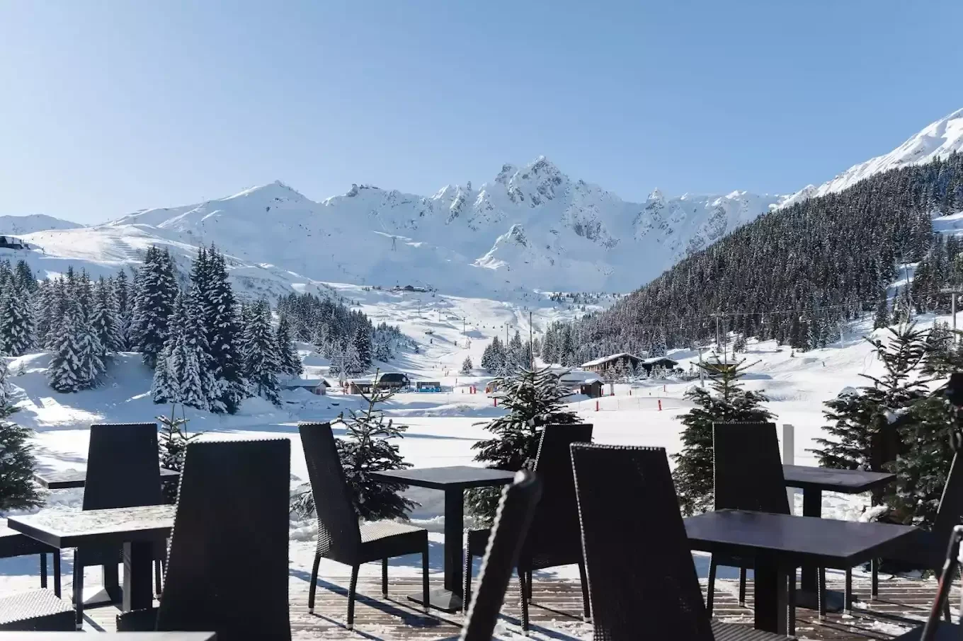 Terrasse extérieure de l’Hôtel Lake Courchevel avec tables et chaises sur la neige. C’est l’endroit idéal pour un déjeuner en plein air avec vue panoramique sur les majestueuses montagnes enneigées.