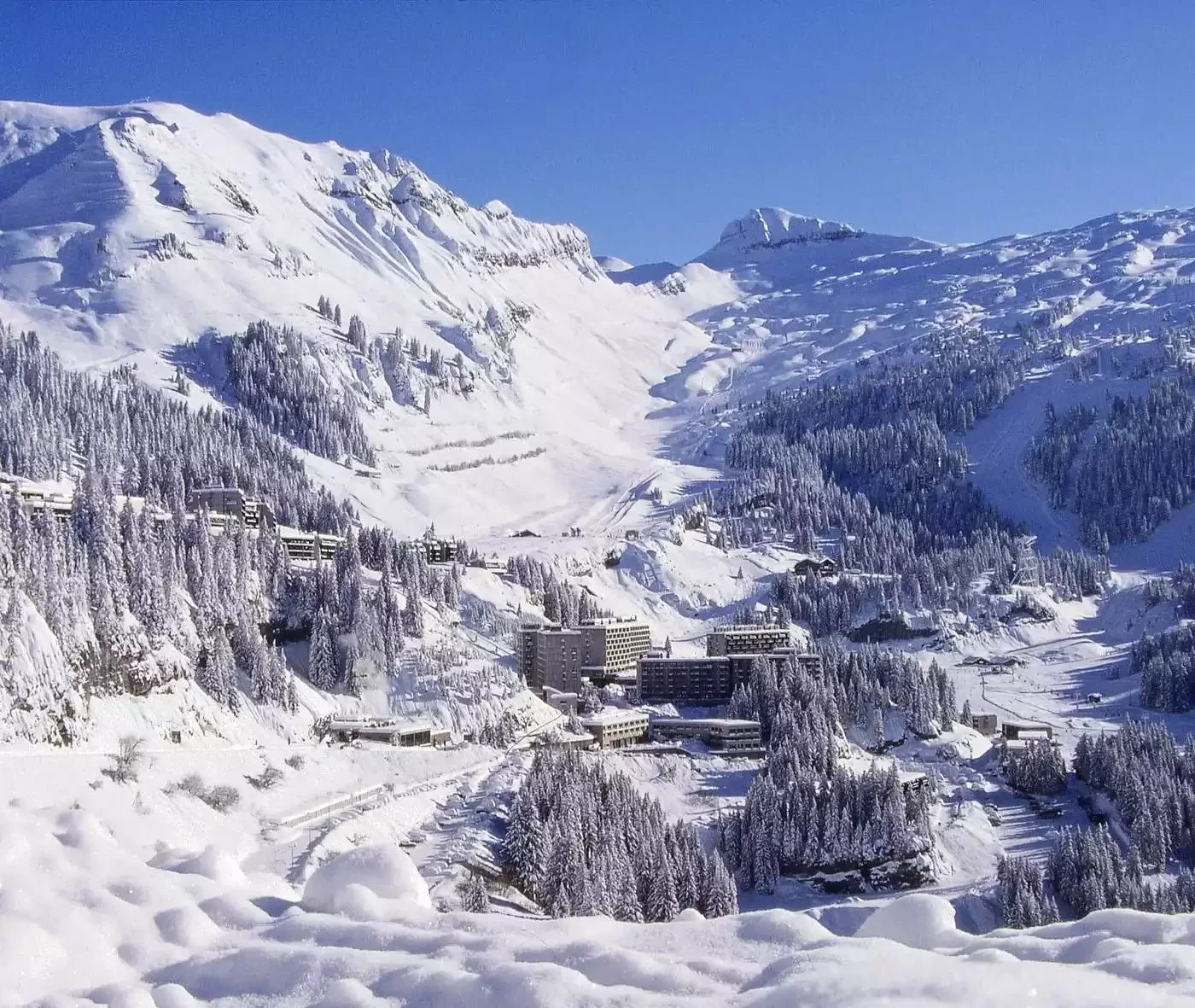 Vue panoramique sur la station de Flaine, avec ses bâtiments de style brutaliste et ses pistes de ski enneigées au milieu des forêts alpines, sous un ciel bleu vif.