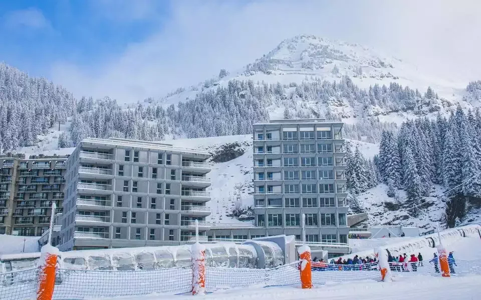 Vue extérieure de l’Alhena & spa hotel, un bâtiment moderne avec façade en béton et grandes fenêtres, situé dans une station de ski enneigée. L’environnement montre des pistes et des montagnes enneigées en arrière-plan.