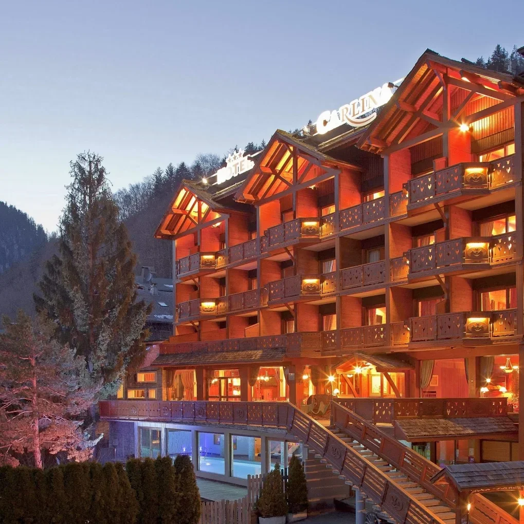Vue nocturne de la façade en bois de l’Hôtel La Carlina à La Clusaz. Les lumières chaudes des chambres et des balcons créent une atmosphère chaleureuse et luxueuse dans la montagne.