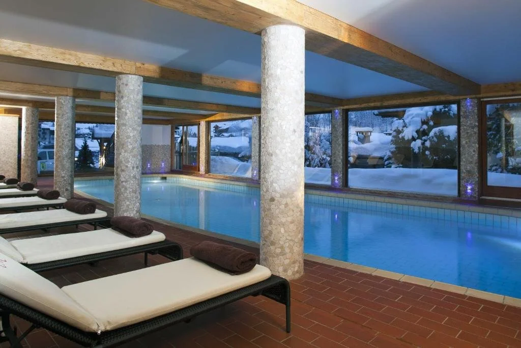 Piscine intérieure de l’Hôtel La Carlina avec de grandes baies vitrées donnant sur un paysage enneigé. L’espace, avec des piliers en pierre et des chaises longues, est parfait pour se détendre après une journée dans la neige.