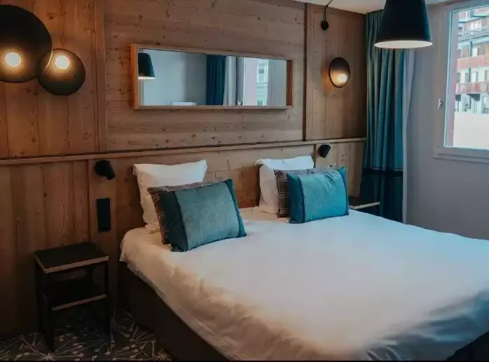Chambre d’hôtel à L’Eden des Cimes avec un design moderne en bois, grand lit et textiles bleus. C’est un espace calme et fonctionnel, idéal pour le repos des participants d’un séminaire Aylo Montagne.