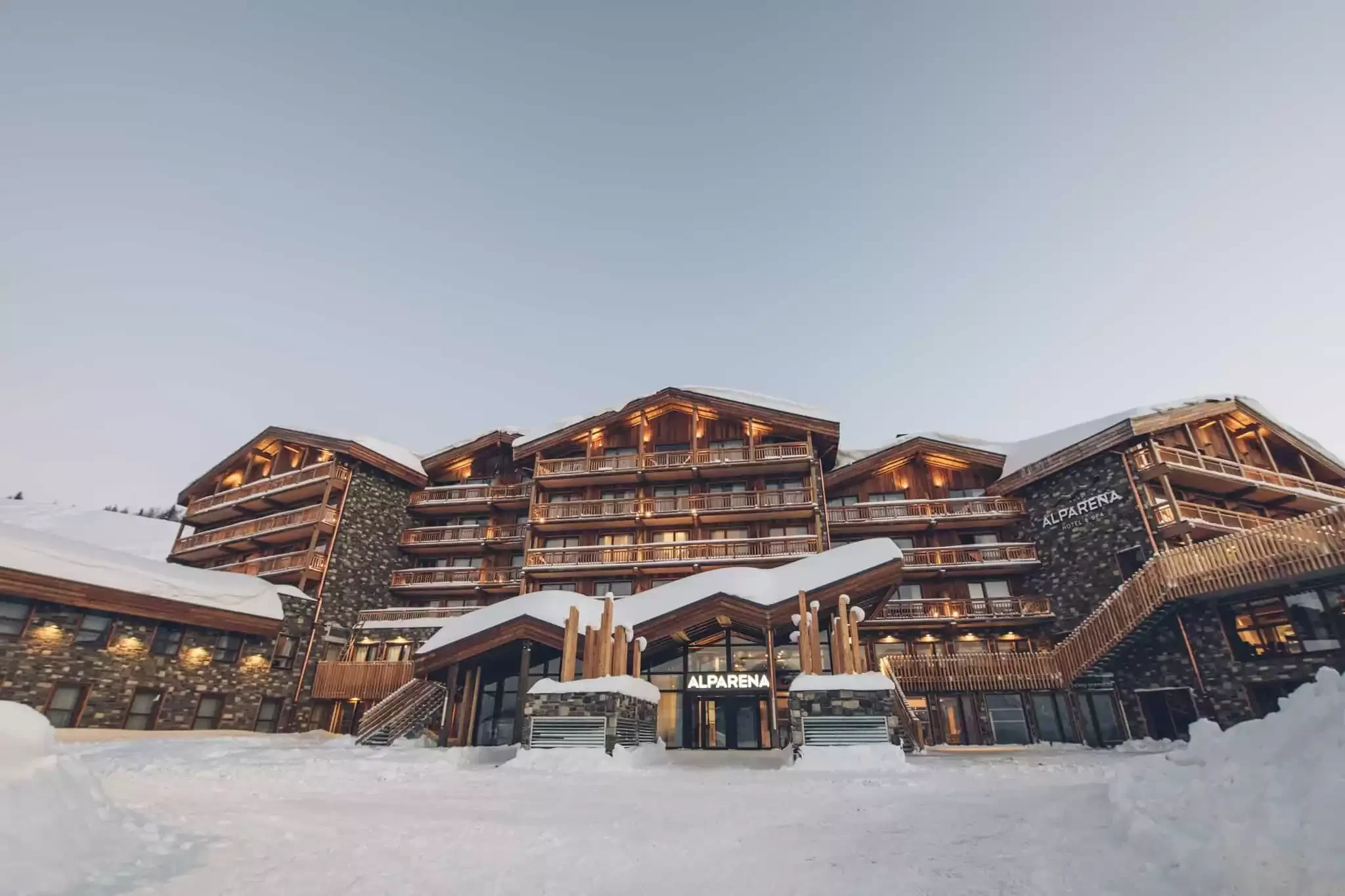 Vue de la façade de l’Hôtel Alparena à La Rosière au crépuscule. Cet hôtel design en bois et pierre, recouvert de neige, est éclairé et crée une atmosphère accueillante.