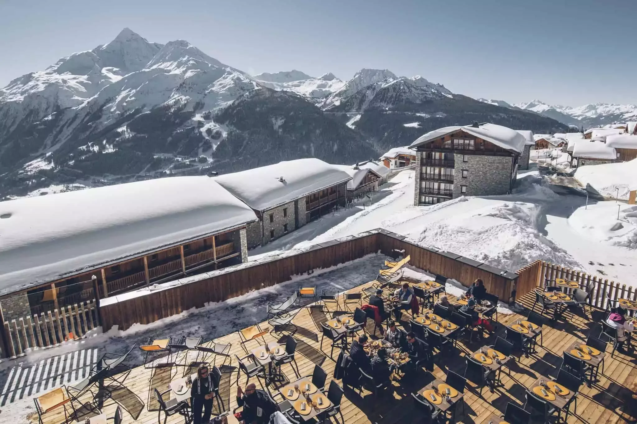 Vue aérienne de la terrasse extérieure de l’Hôtel Alparena, avec tables et chaises en bois sur la neige, avec vue panoramique sur les montagnes et la station de ski.