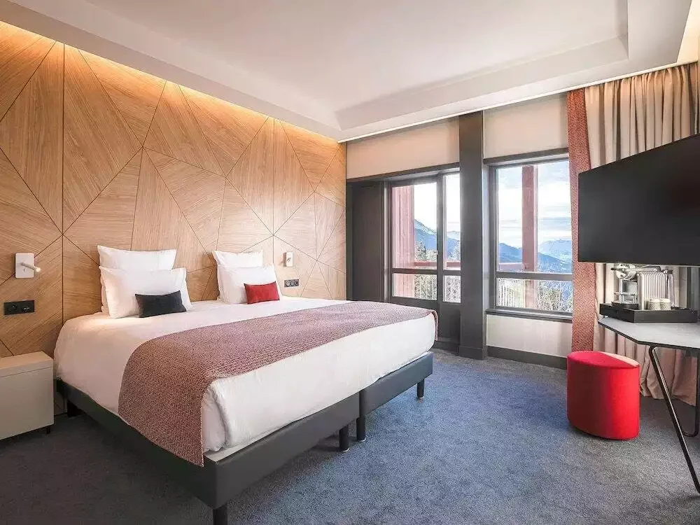 Chambre design à l’Altezza arc 1800 4* avec des murs en bois, un grand lit avec une couette rouge et une télévision à écran plat, offrant une vue sur la montagne.