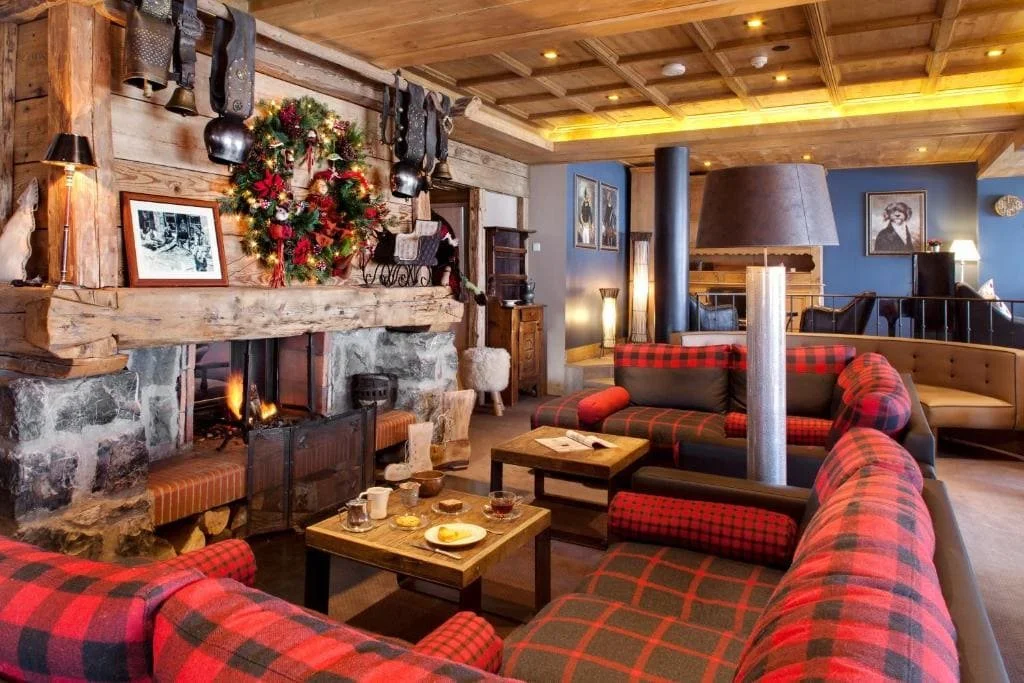 Salon de La Marmotte Hôtel avec une grande cheminée en pierre et des canapés à carreaux rouges. Cet espace cosy est parfait pour la cohésion d'équipe et les moments de détente d'un séminaire d'entreprise Aylo Montagne.