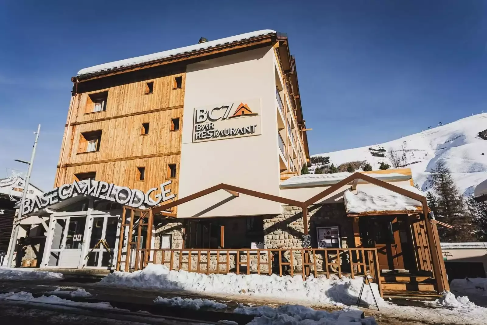 façade extérieure du Base Camp Lodge avec son entrée couverte de neige. L’hôtel a une architecture moderne qui combine des murs en bois et des tons clairs, créant une atmosphère accueillante dans la montagne.