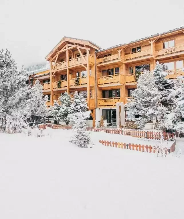 Vue extérieure du Chalet Mounier, un hôtel de style chalet en bois avec balcons et toits enneigés. Le bâtiment est entouré d’arbres enneigés, créant une atmosphère accueillante et festive en hiver.