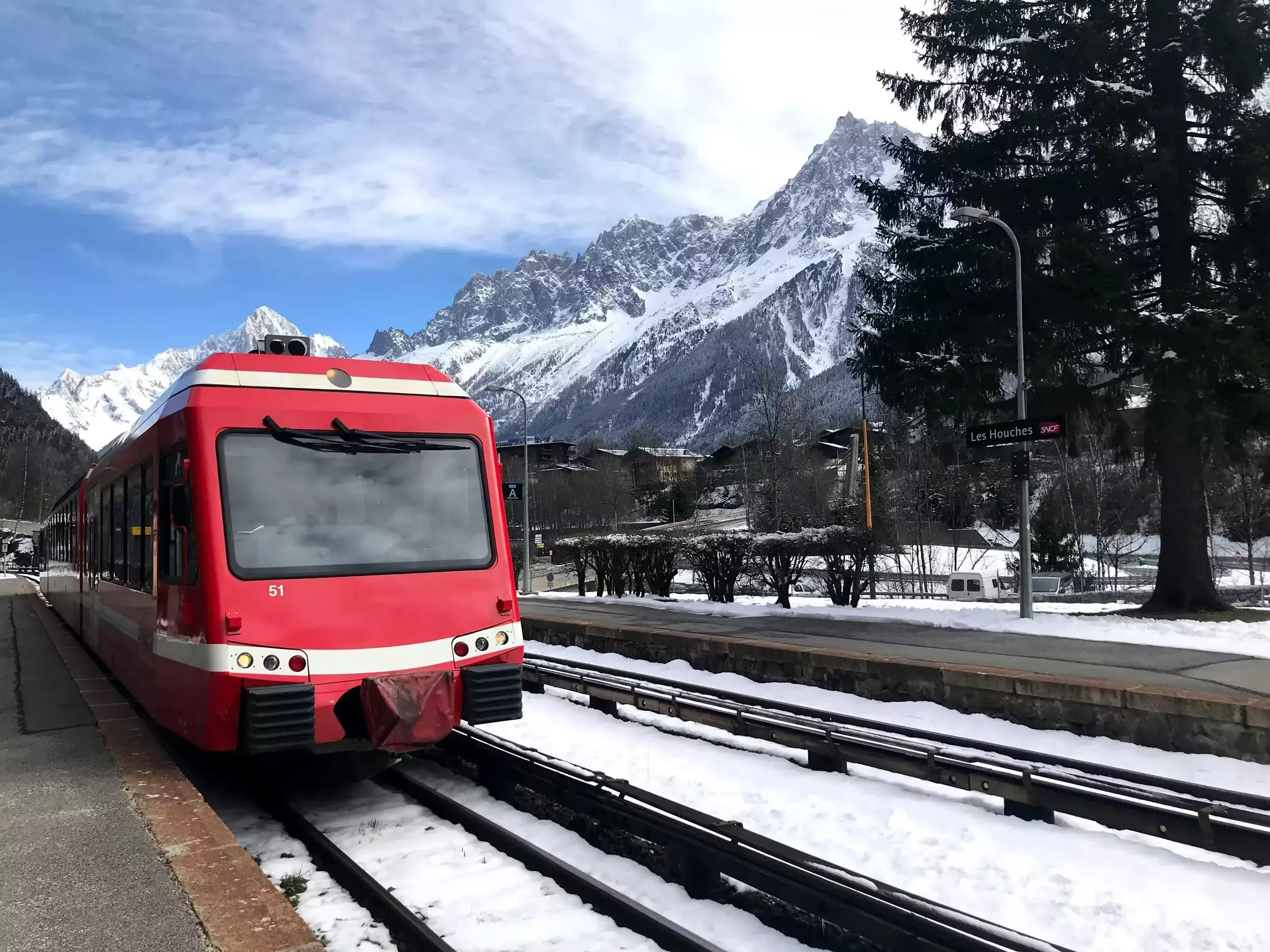 Un train rouge de montagne se trouve à la gare des Houches, avec de la neige sur les rails. En arrière-plan, de majestueuses montagnes enneigées s’élèvent et des chalets traditionnels sont appréciés.